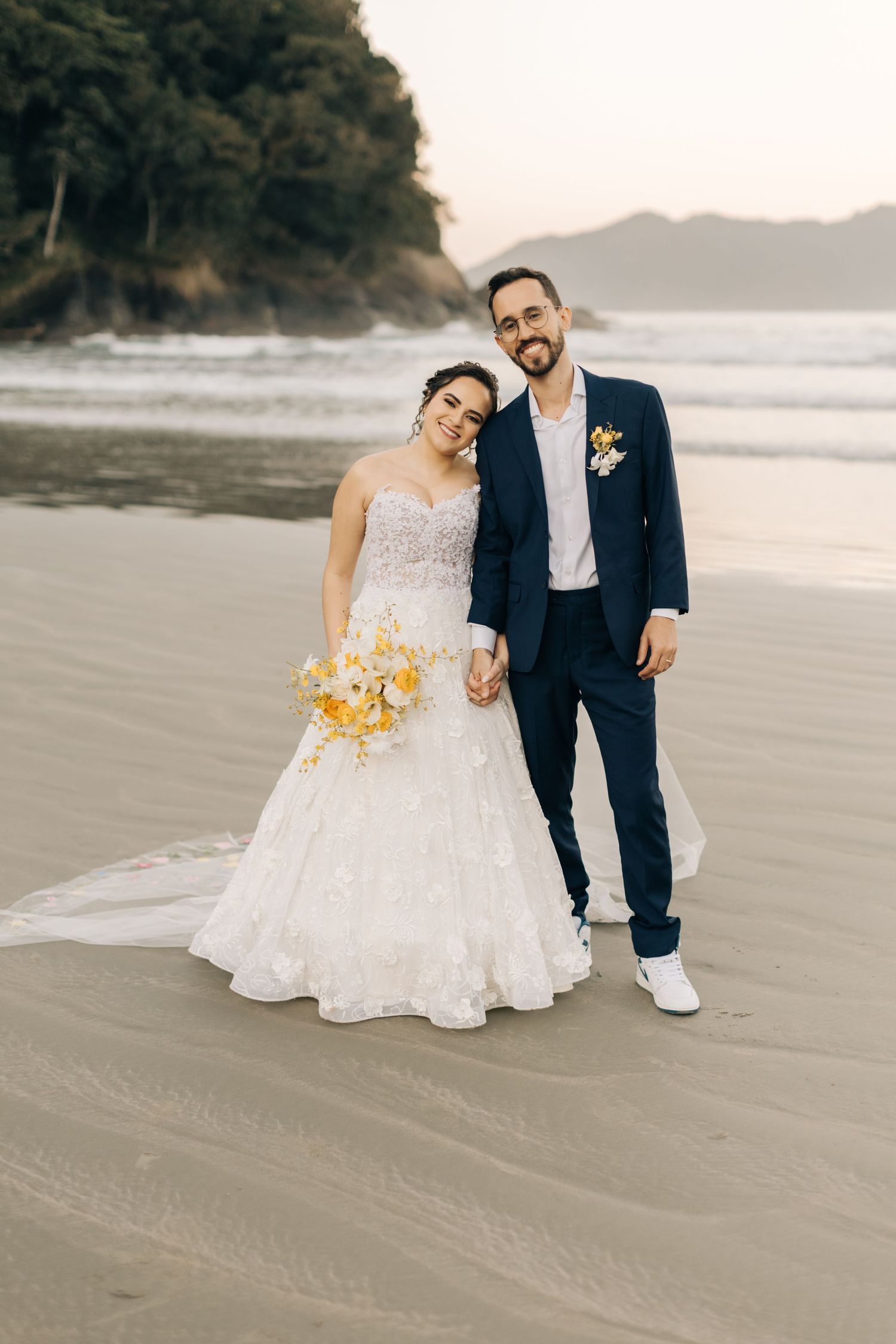 A wedding couple poses on a scenic beach with mountains in the background as the bride holds a yellow and white bouquet.
