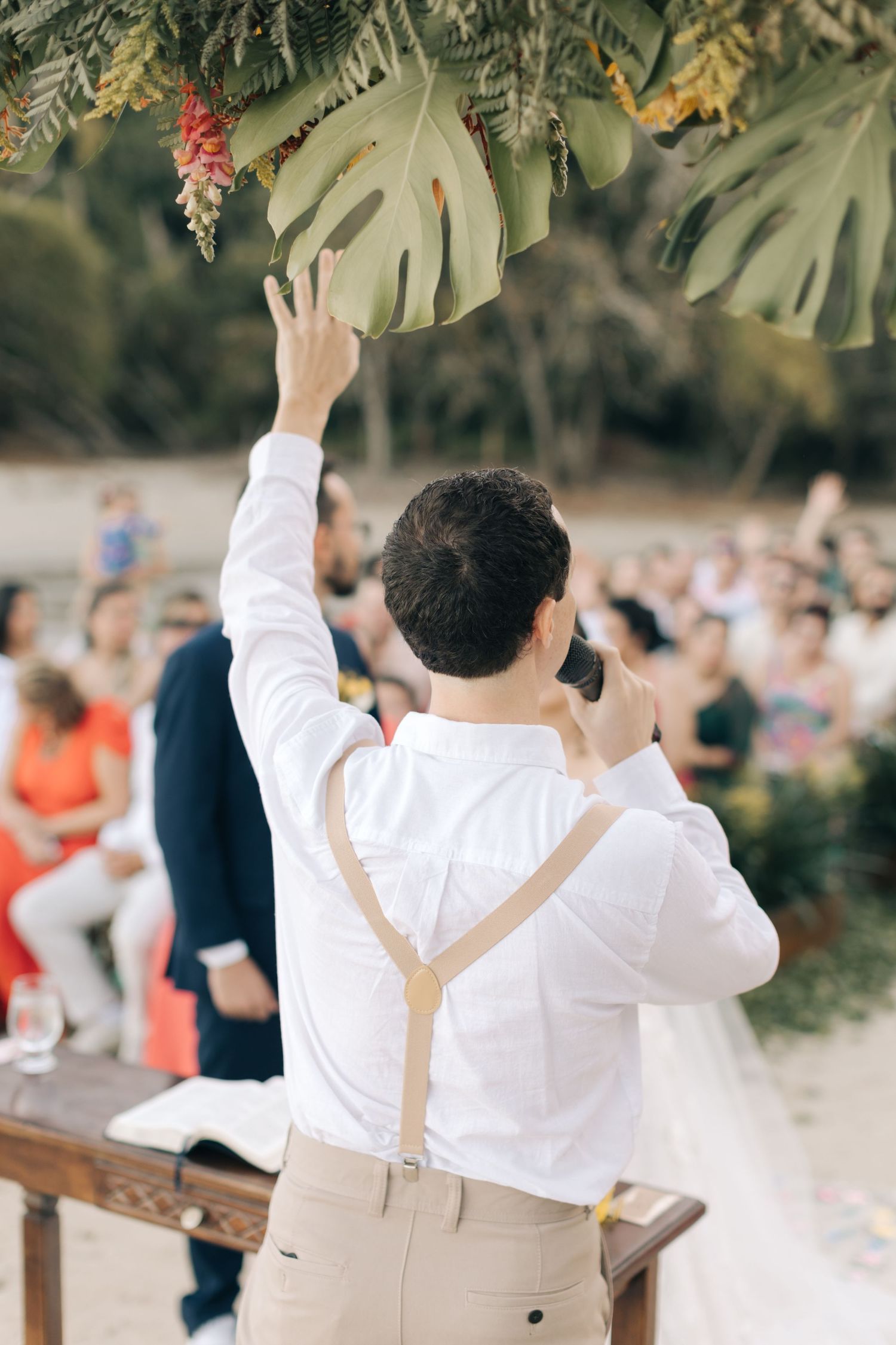 Wedding guest in suspenders gestures enthusiastically during an outdoor celebration.
