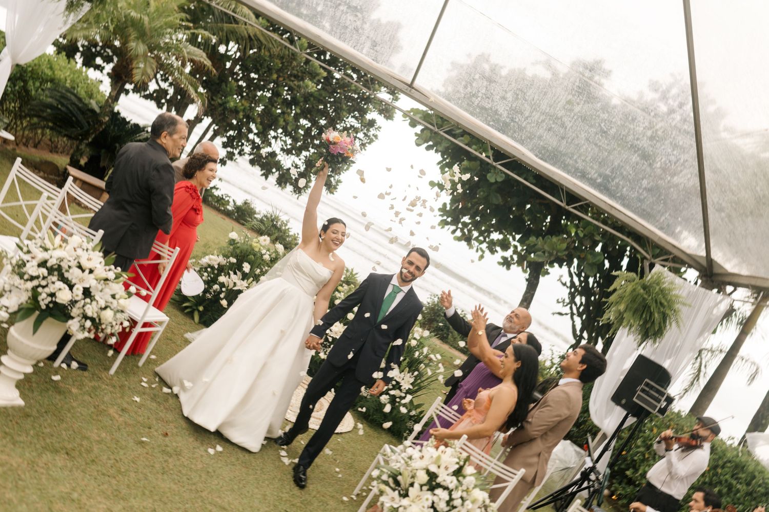 Wedding guests celebrate during an outdoor ceremony under a white canopy with floral arrangements.