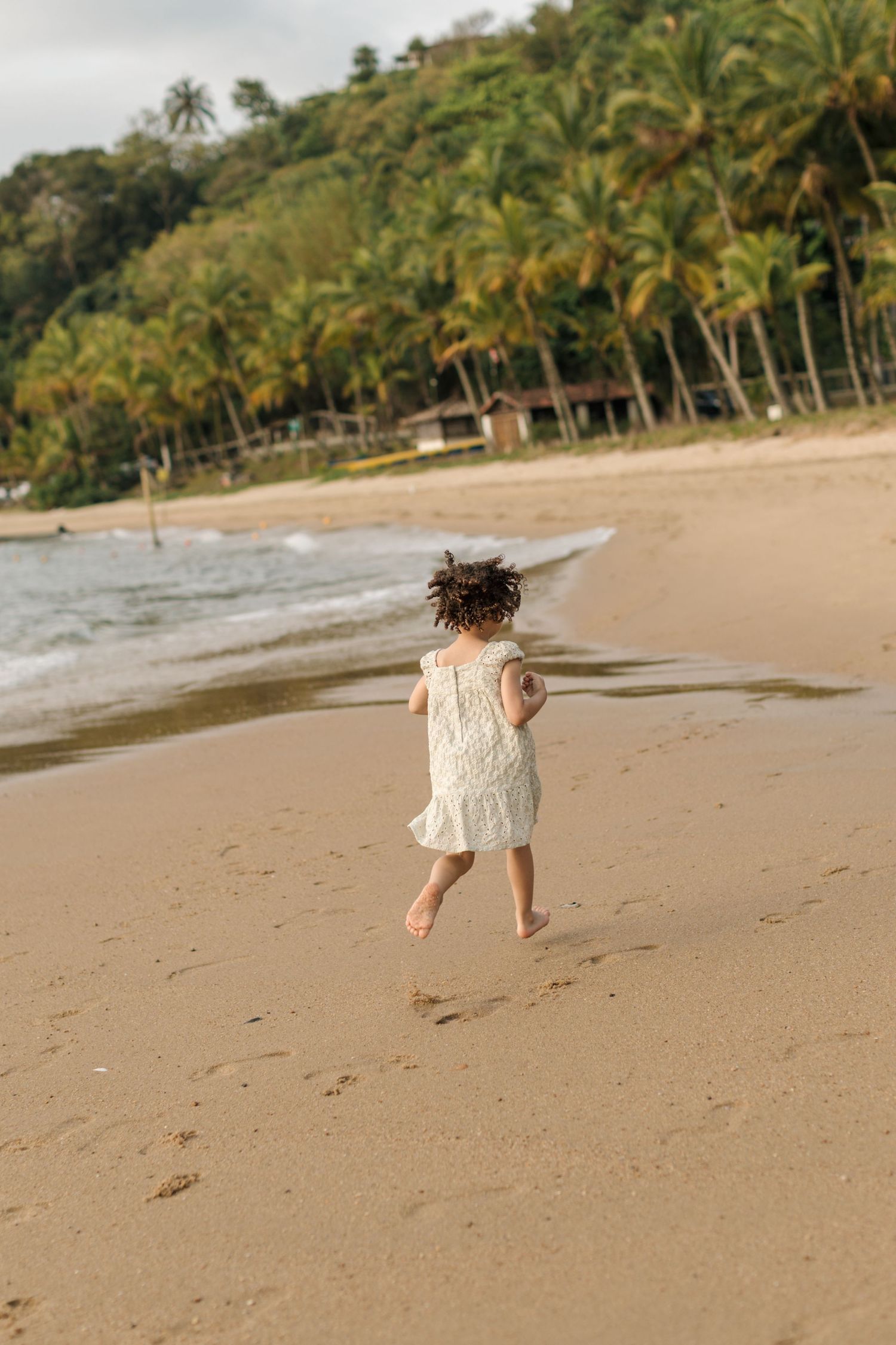 Child in white dress runs joyfully along a tropical beach lined with palm trees.