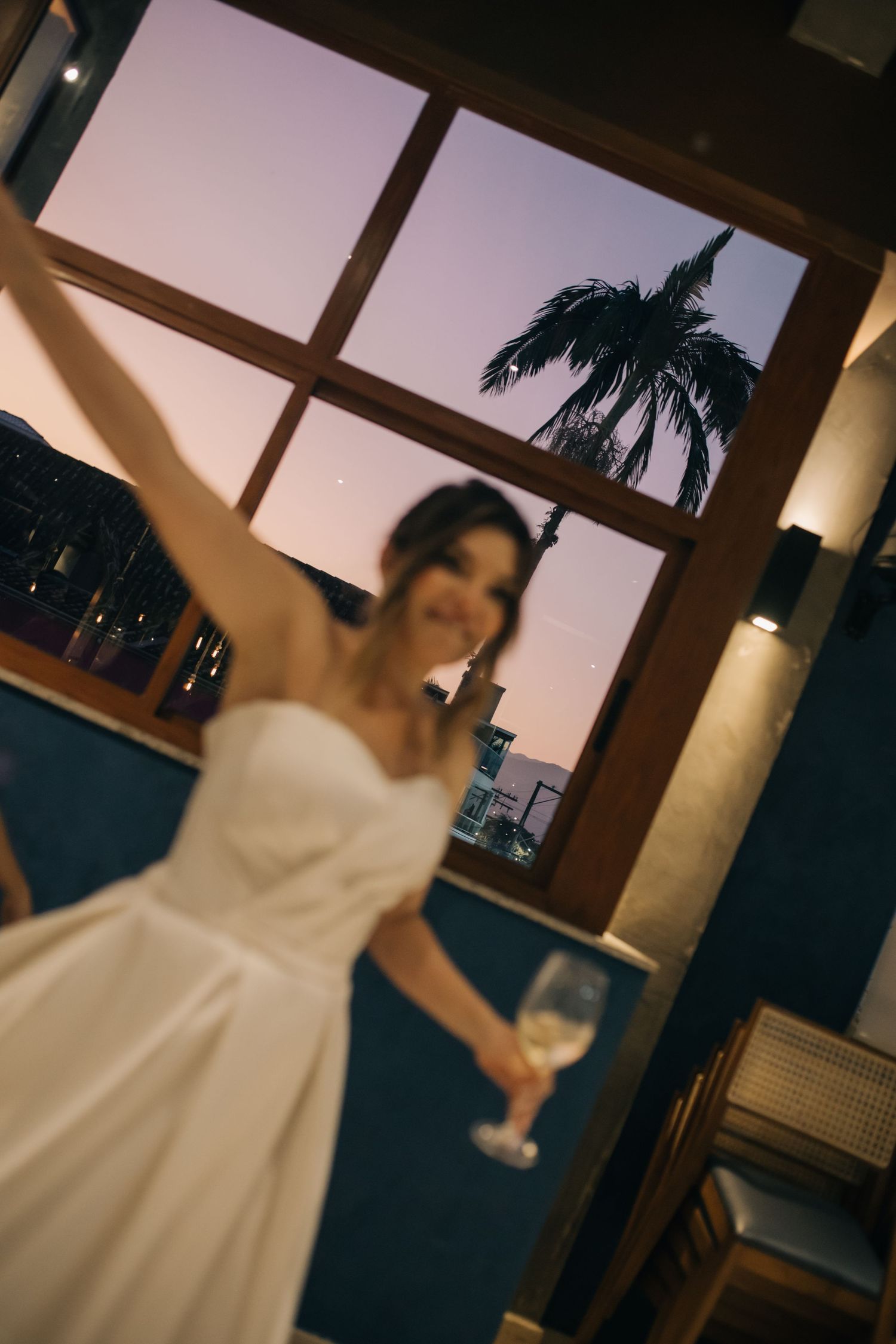 White wedding dress silhouetted against a window with palm tree view.