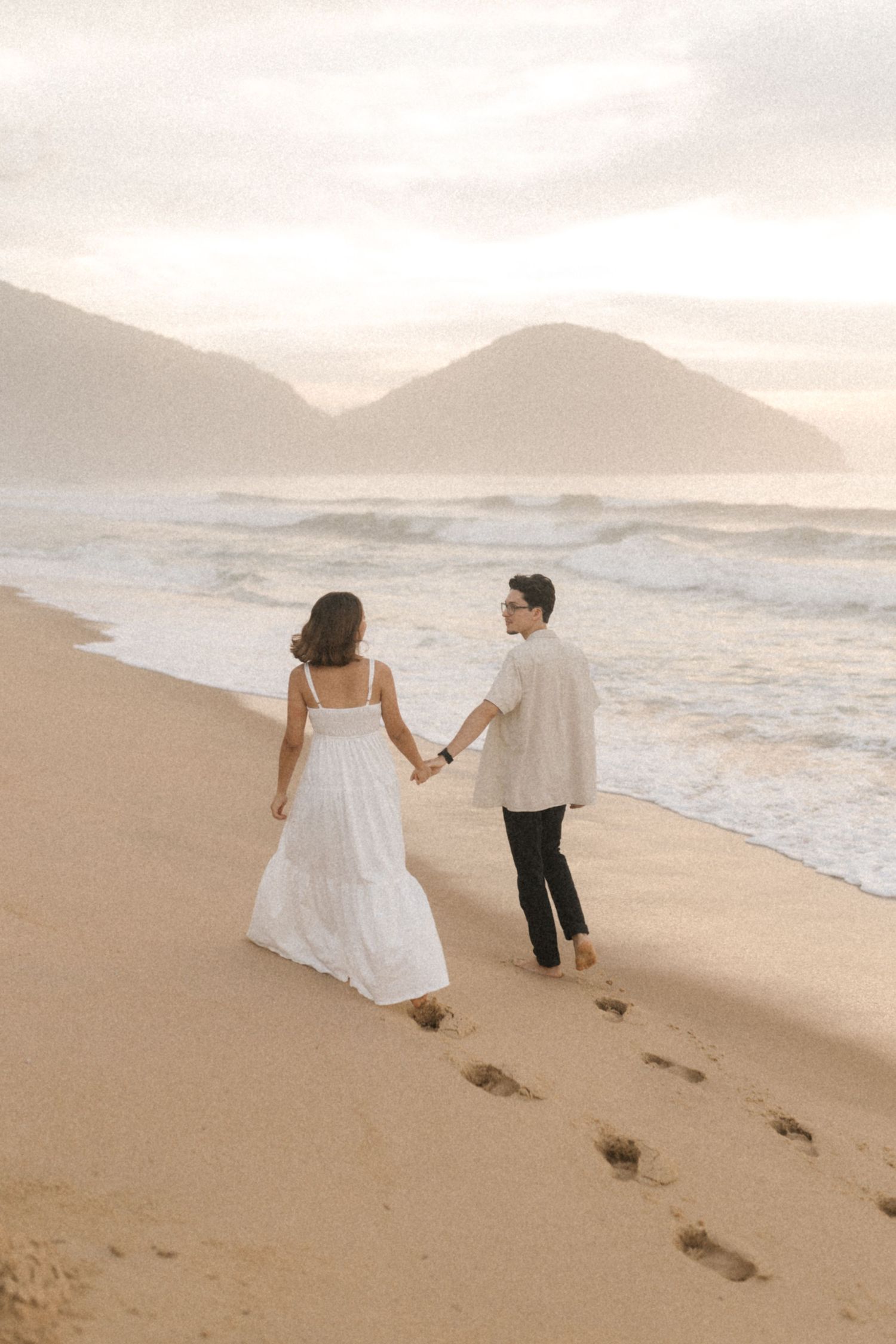 Couple walking hand in hand along a scenic beach shore with mountains in the background at sunset.