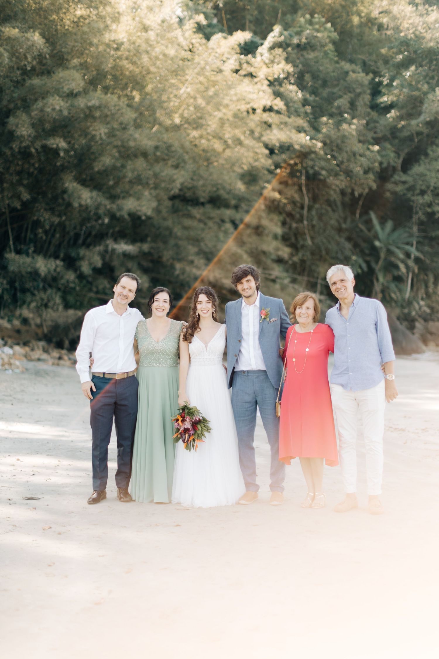 Wedding party in pastel dresses and suits poses together on a sandy beach with trees in the background.