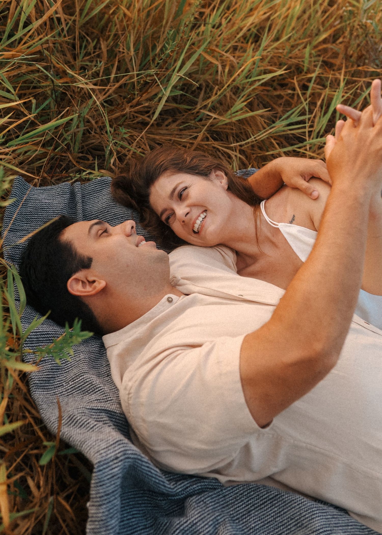 Two people lie together on a blanket in tall grass during an outdoor photo session.