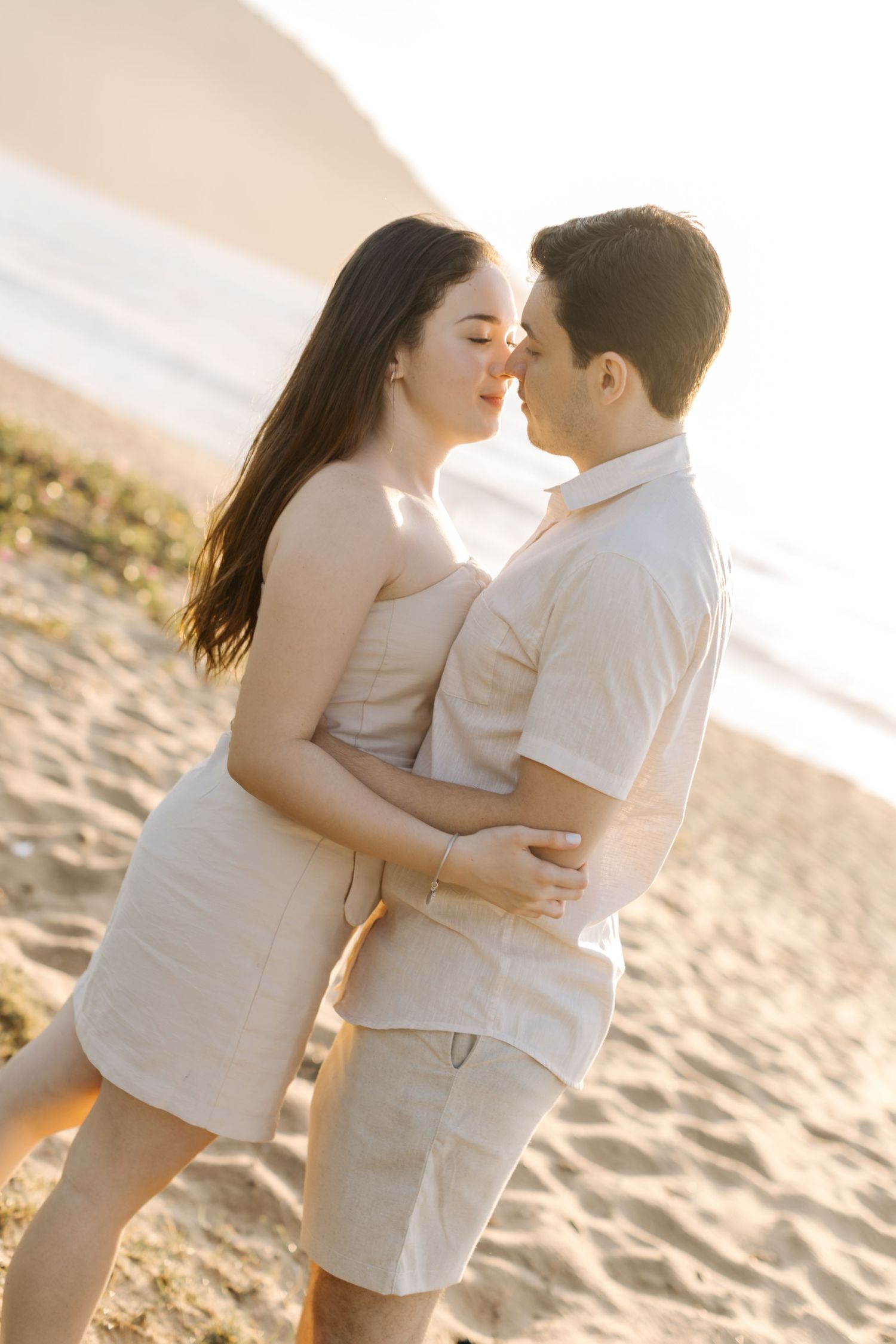 Romantic couple embracing on sandy beach at sunset in matching light colored outfits.
