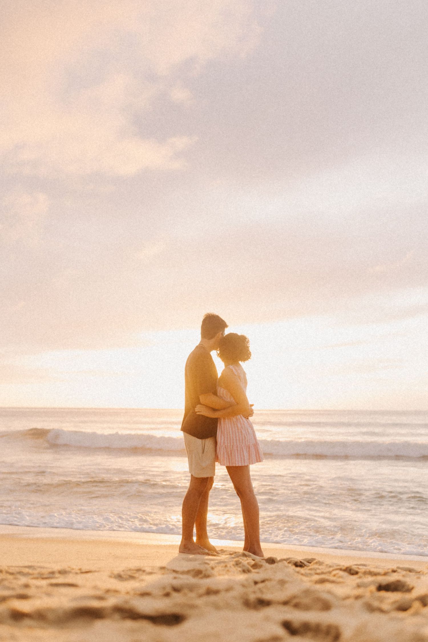 Casal em silhueta se abraça na praia ao pôr do sol com ondas do mar e céu rosa no fundo.