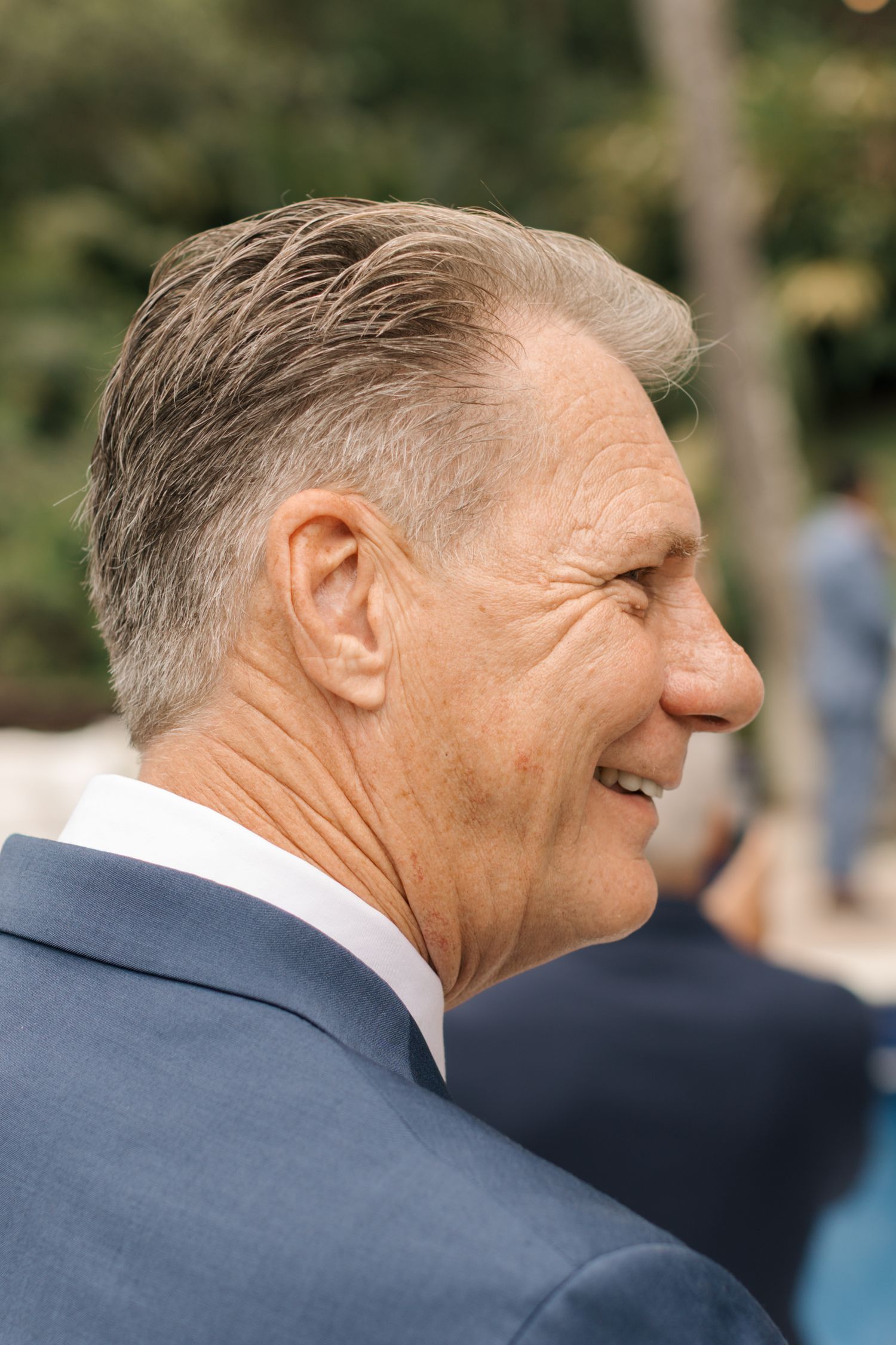Side profile view of someone in a navy suit laughing joyfully during an outdoor event.