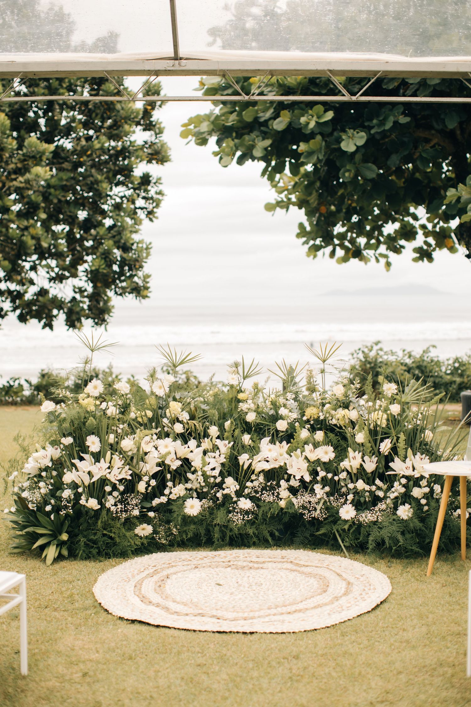 Garden wedding backdrop with lush white flower arrangements and circular rug on lawn.