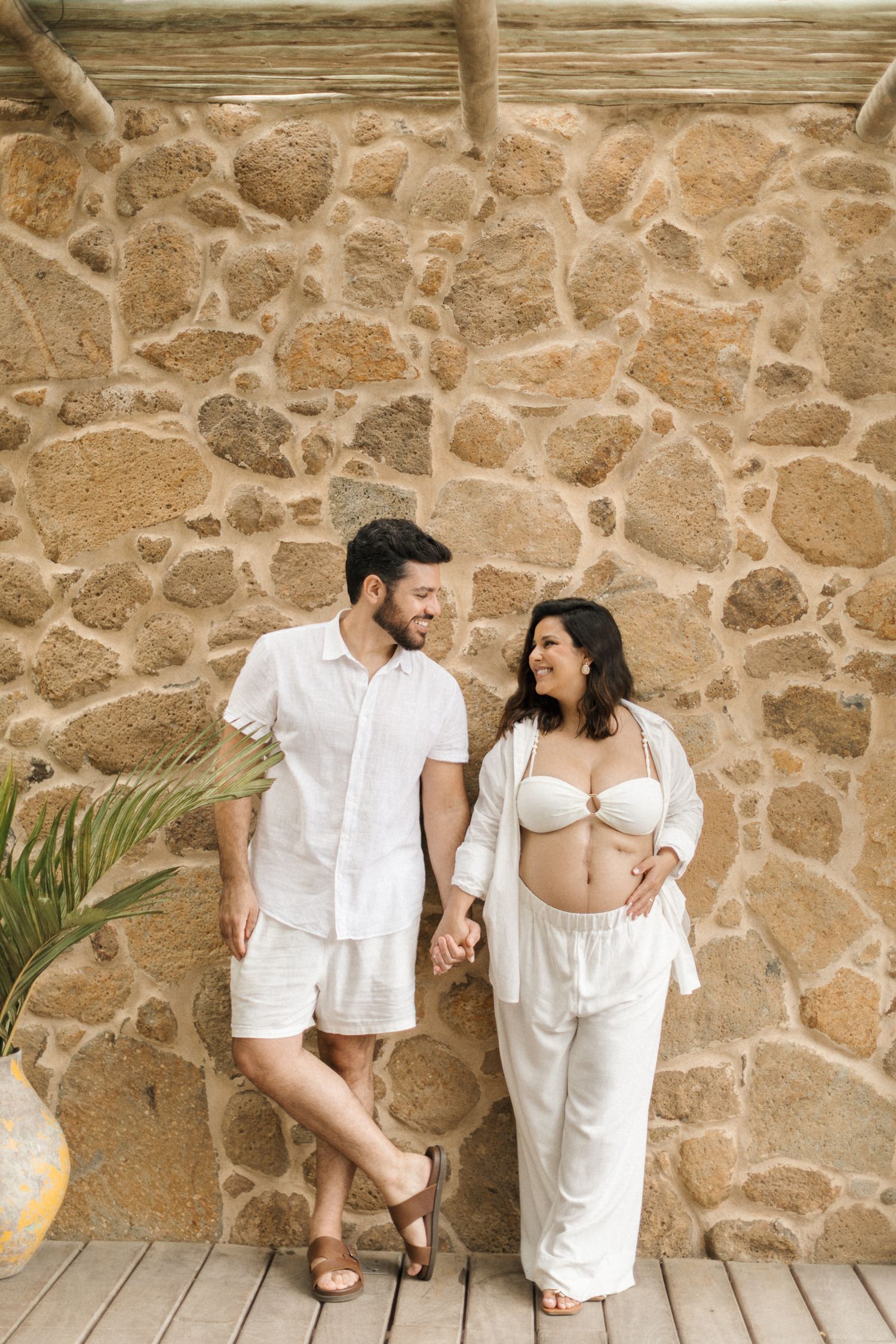 Couple in white summer outfits holding hands and smiling against rustic stone wall with palm frond.