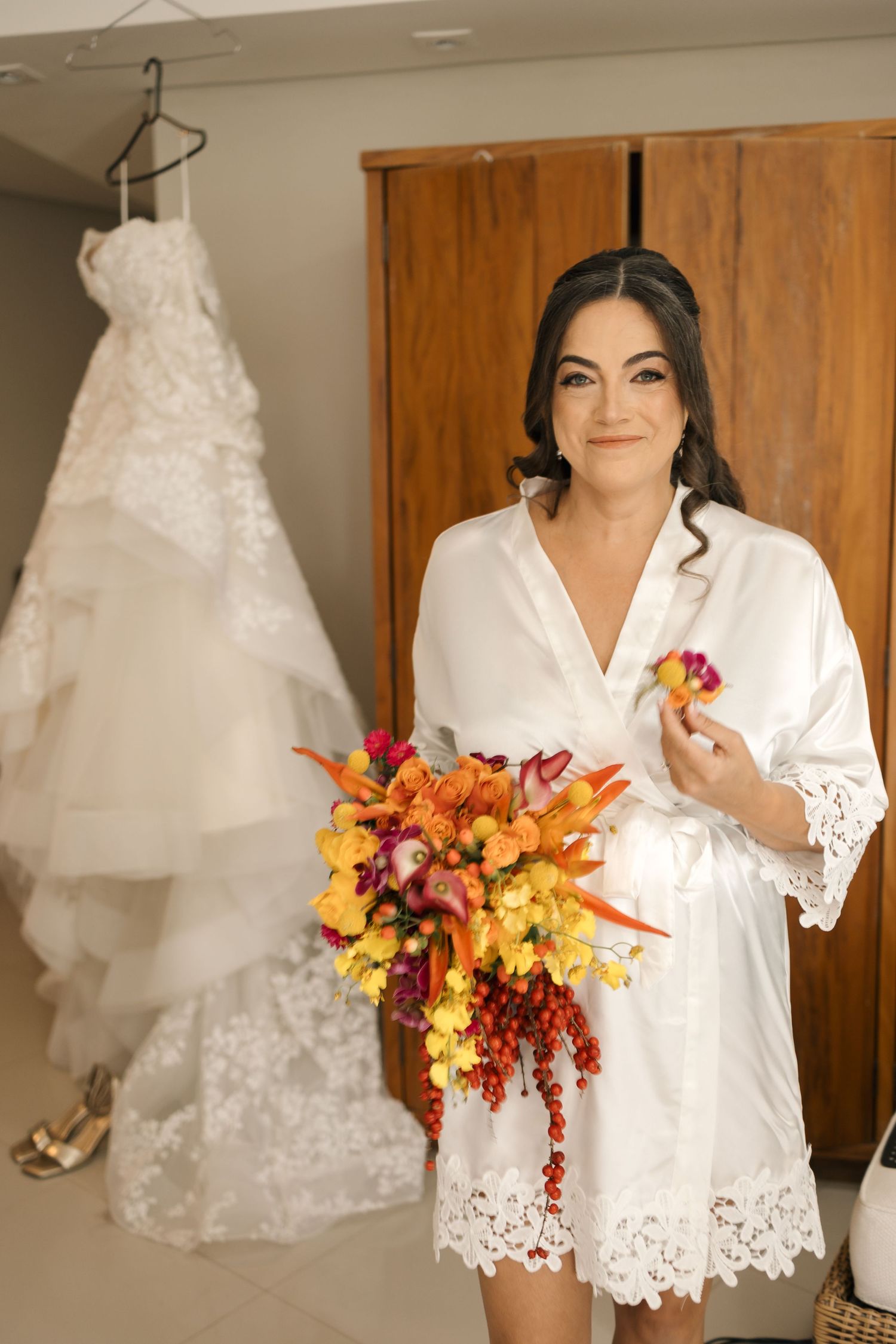 A white lace wedding robe hangs near a wedding dress, with autumn-colored flowers in the foreground.