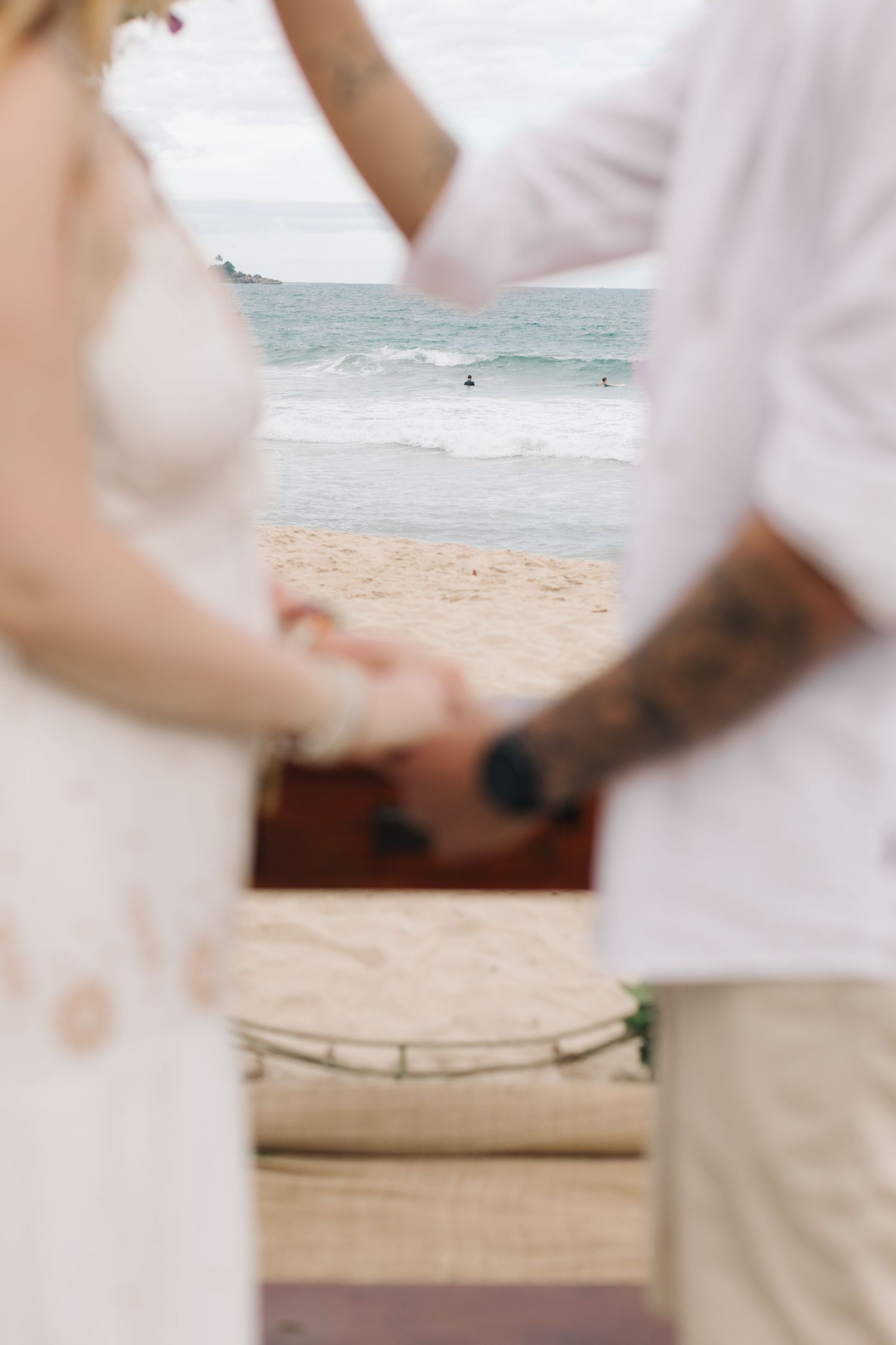 A close-up view of hands holding each other on a sandy beach with ocean waves in the background.
