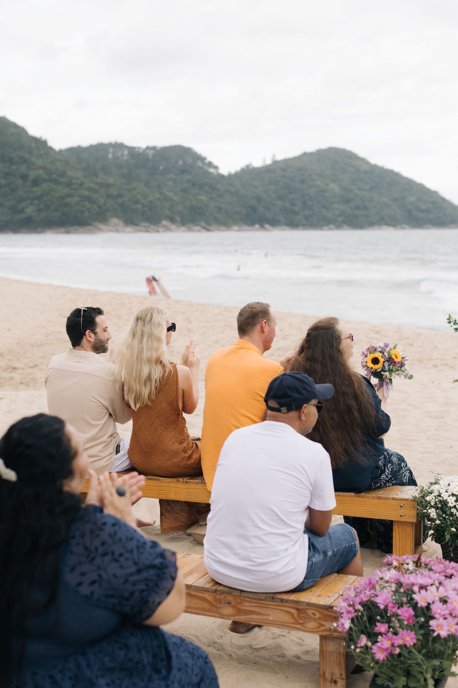 Wedding guests seated on wooden benches at a beach ceremony with mountains in the background.