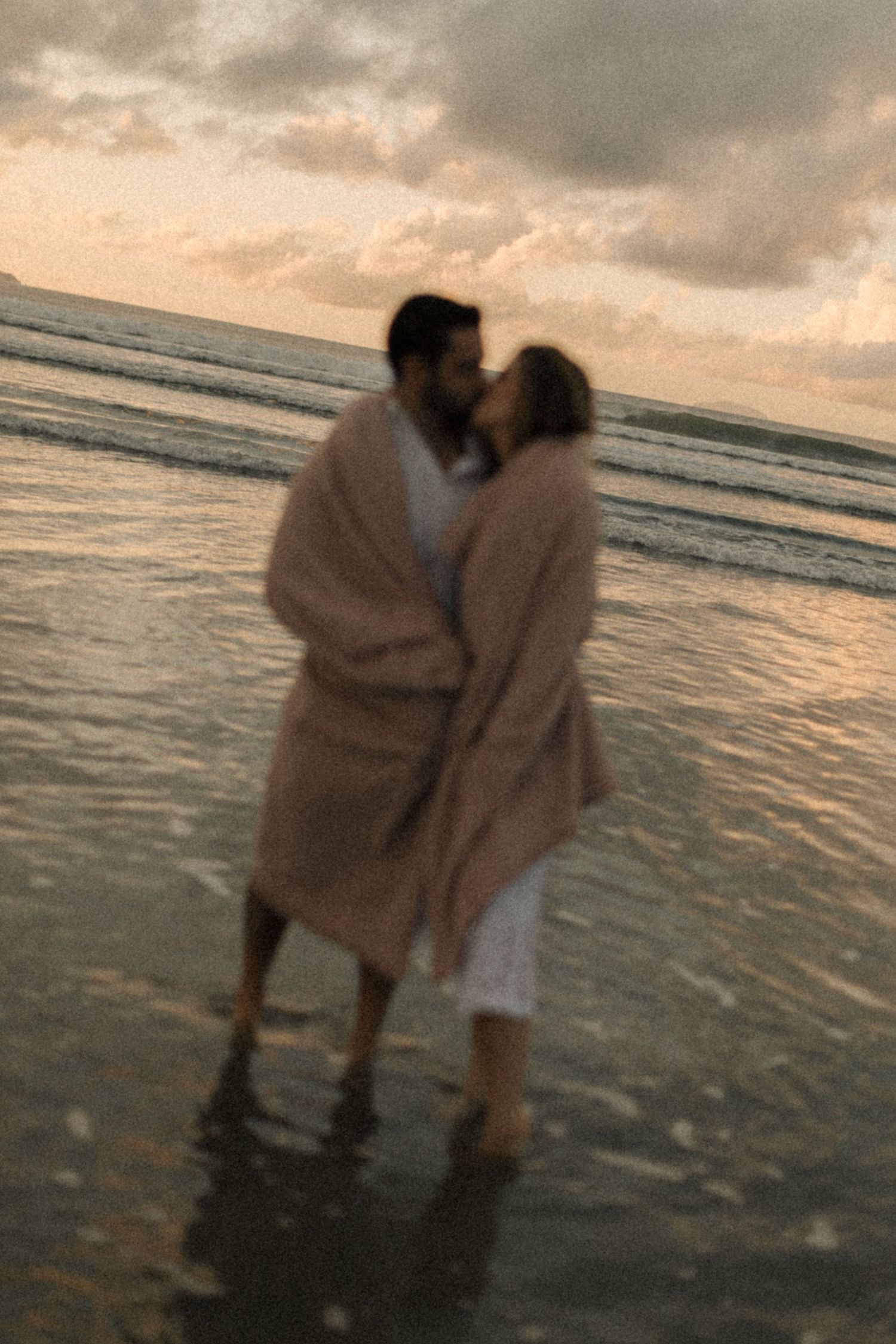 A romantic couple wrapped in a pink blanket shares an intimate moment on the beach at sunset.