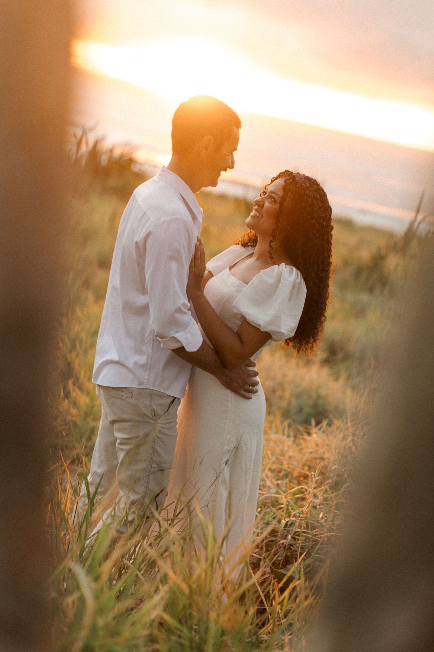 A romantic couple embraces in a golden wheat field during sunset, wearing white outfits for their engagement photos.