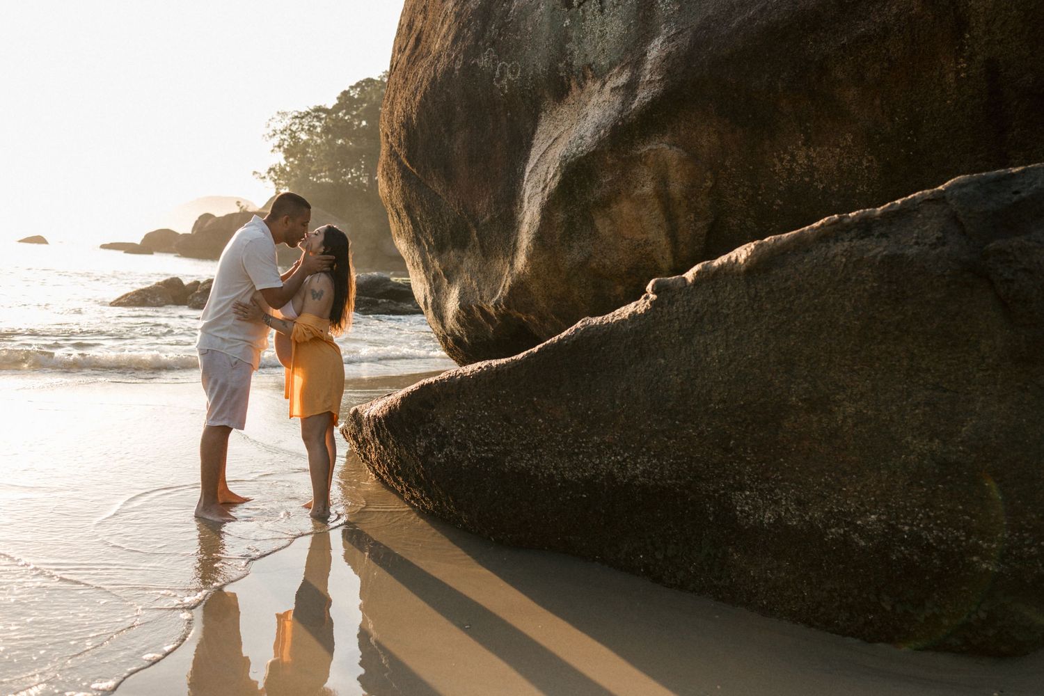 Couple shares intimate moment by large rocks on beach during sunset.