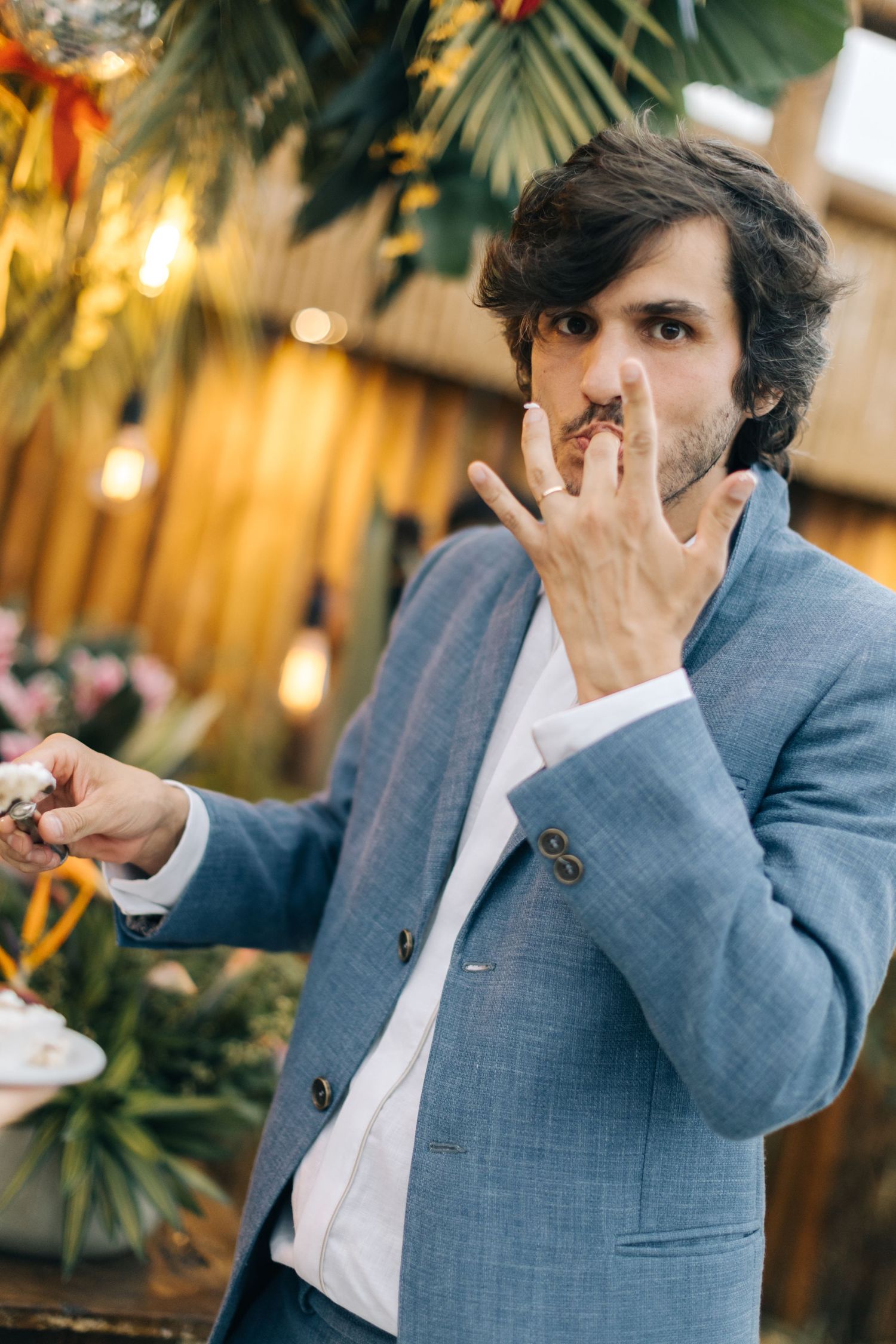 Wedding guest in blue suit tastes dessert at tropical-themed reception with wooden backdrop.