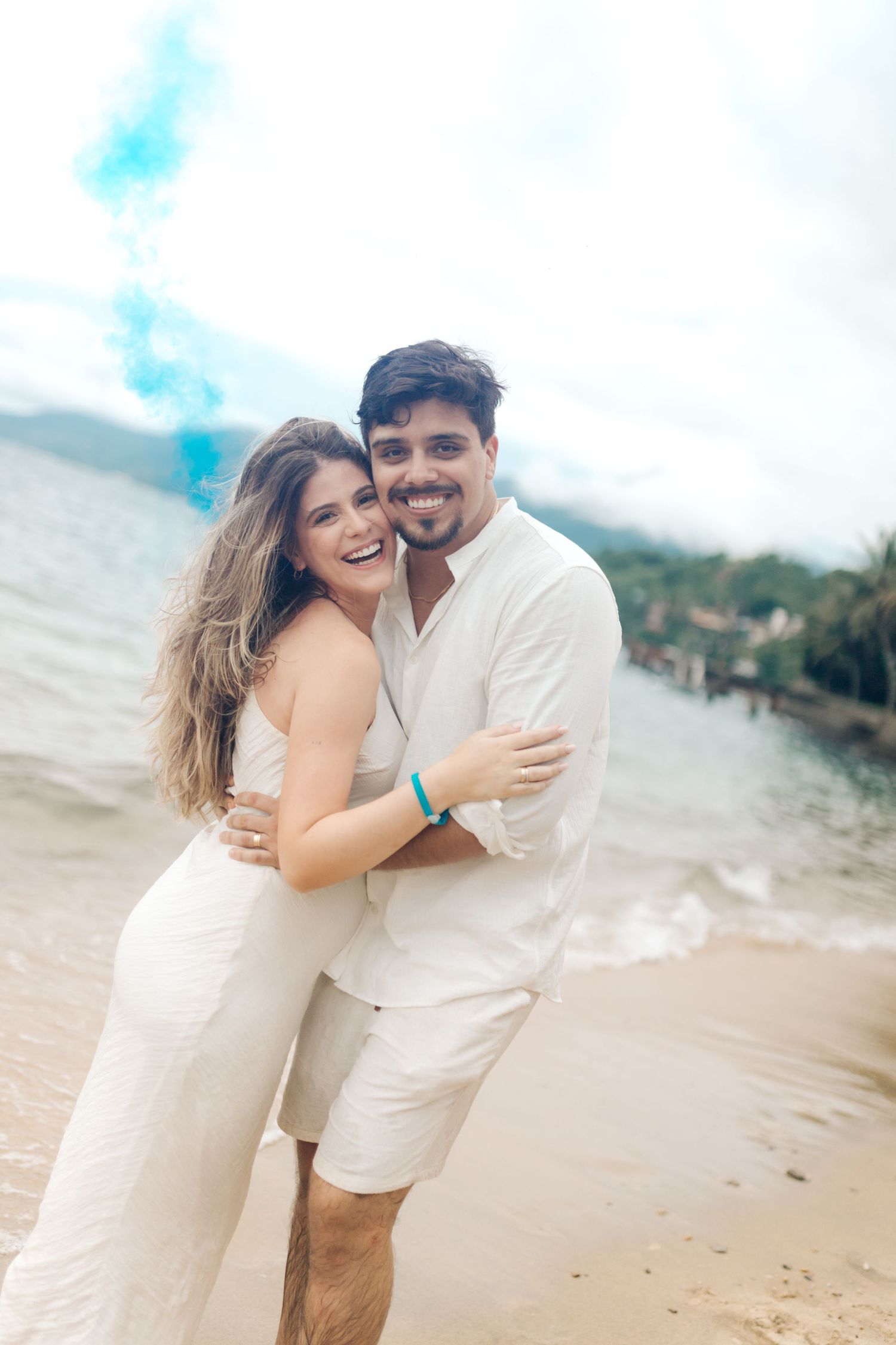 A couple in white outfits embraces and smiles on a tropical beach with blue smoke effects in the background.
