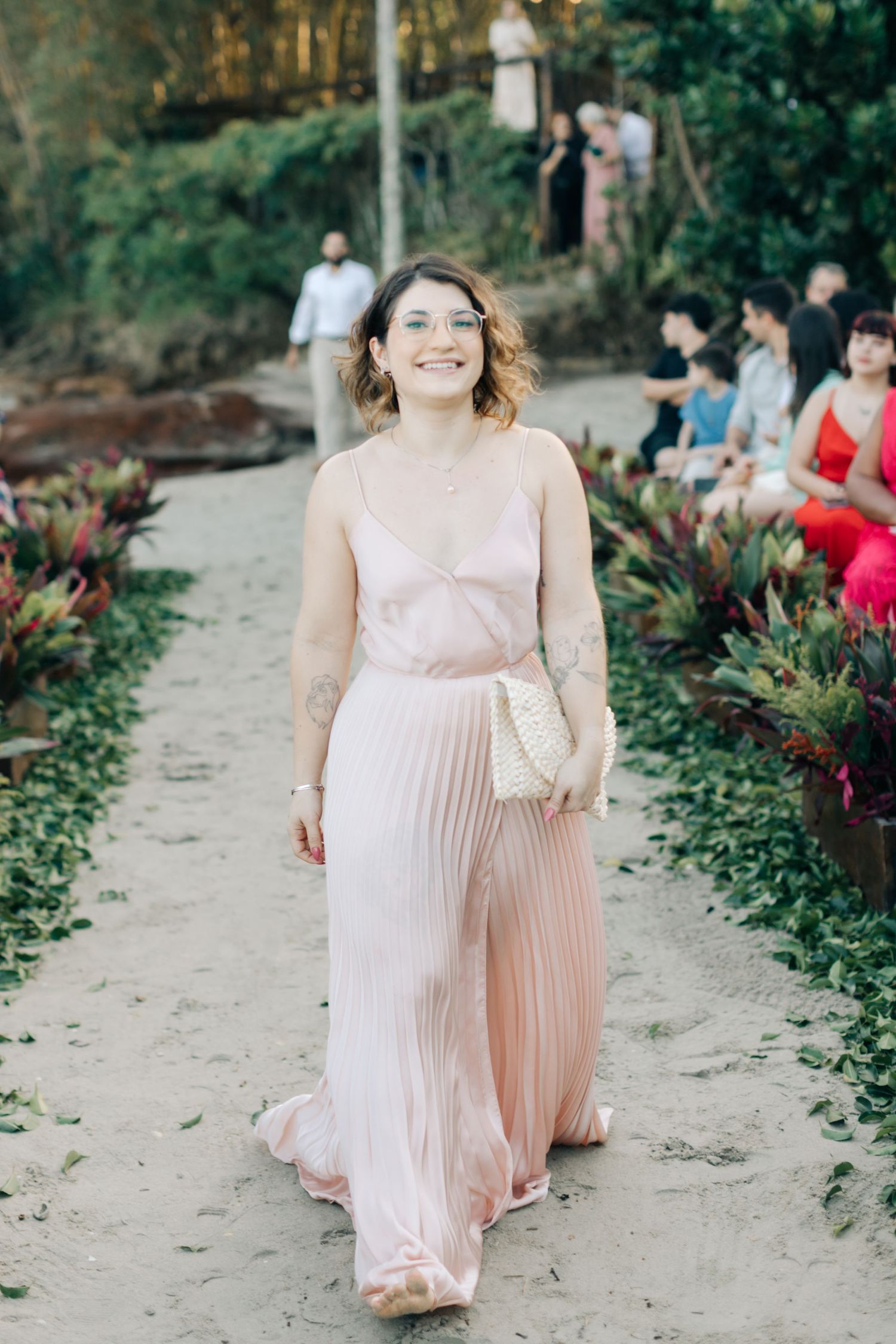 Person in a flowing blush pink dress walking down a sandy aisle at an outdoor ceremony.