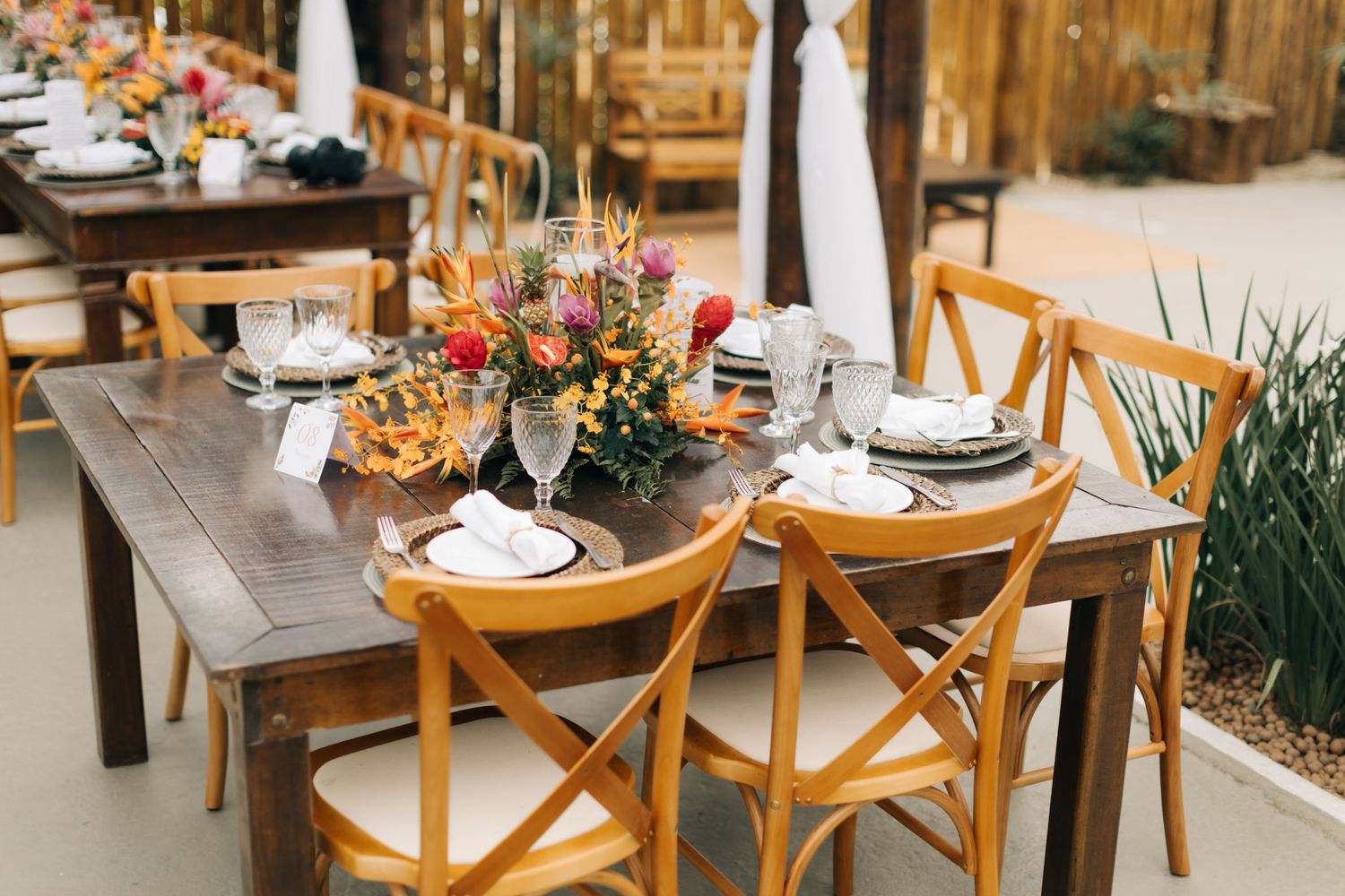 Rustic outdoor wedding table setting with wooden chairs and tropical floral centerpieces.