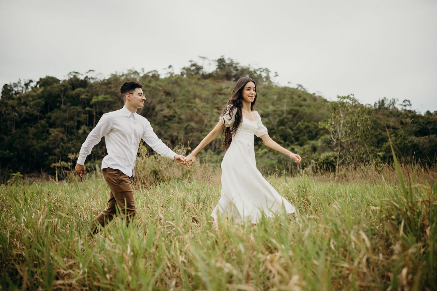 A couple holding hands while walking through tall grass on a hillside.