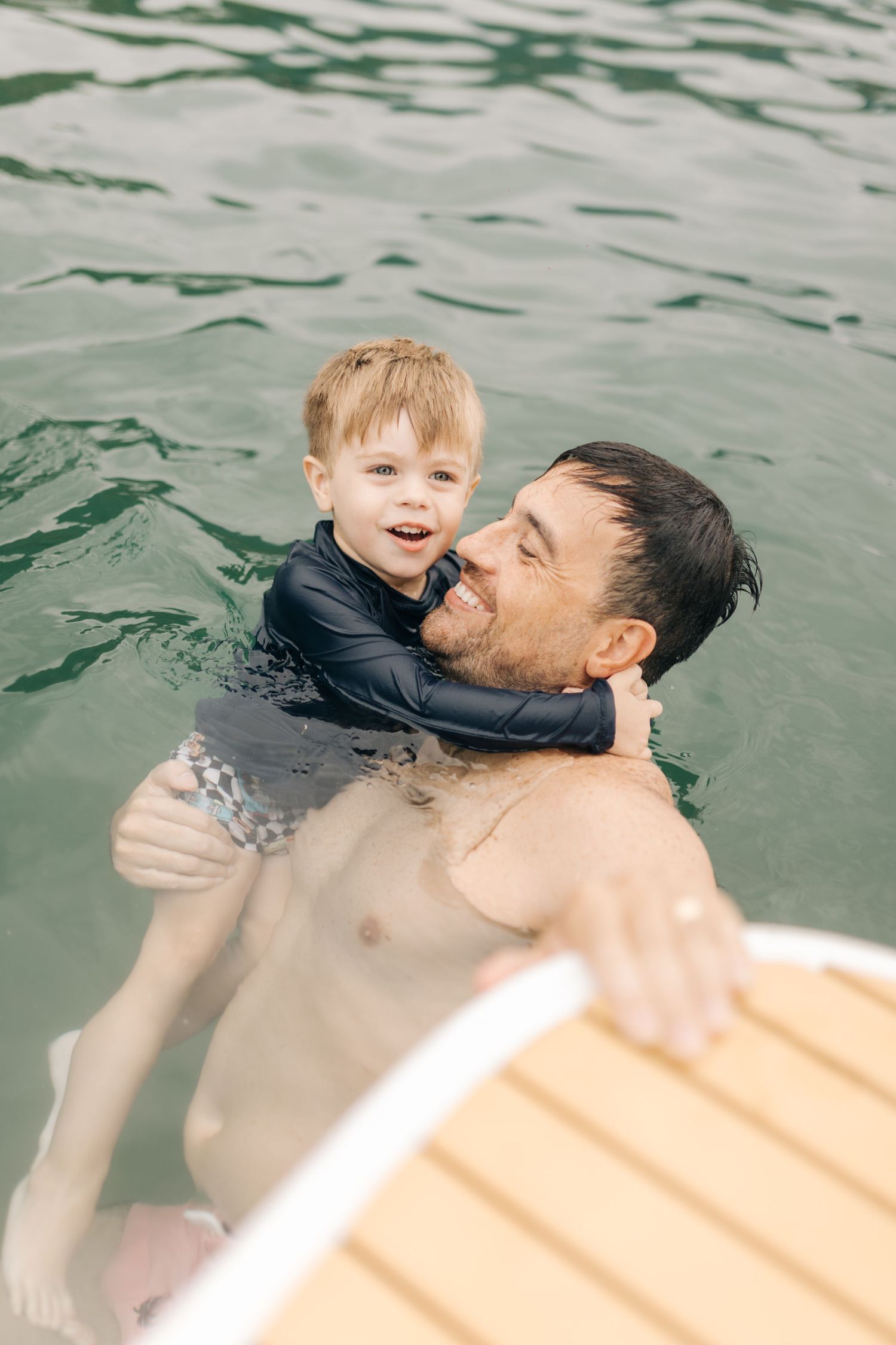 Father and young child enjoying time together swimming in green water near wooden dock.