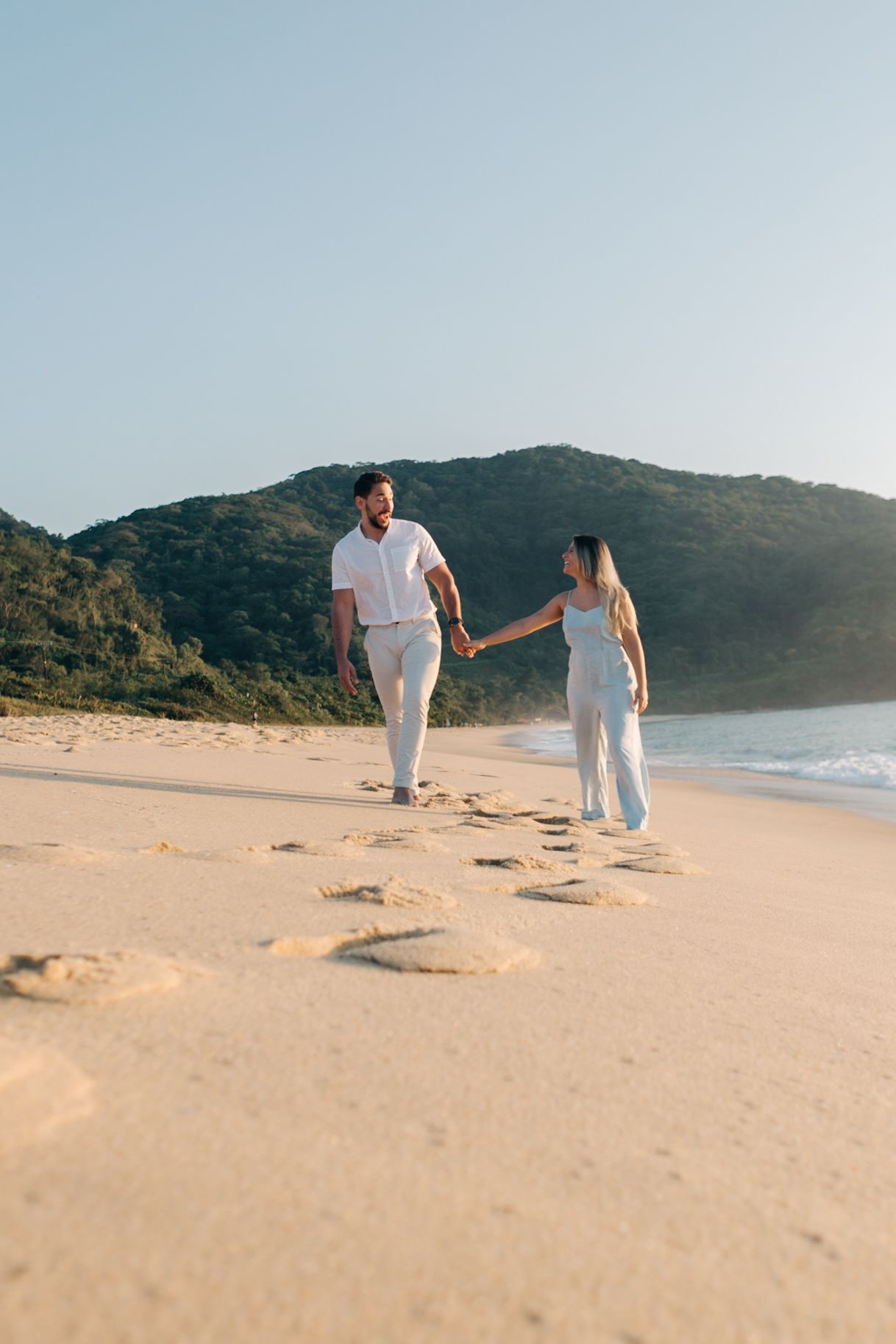 Couple in white outfits walking hand in hand on a scenic beach with mountains in the background.