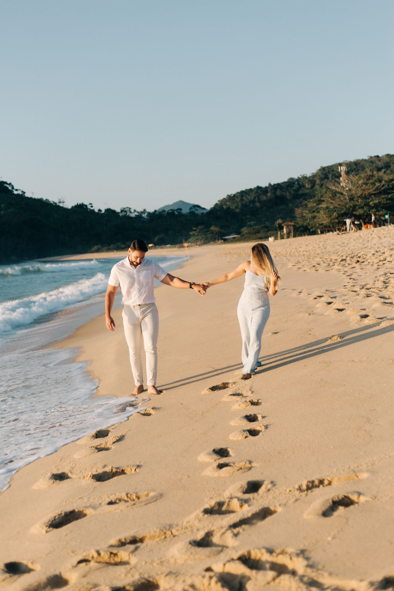 Two people in white outfits holding hands while walking on a sandy beach, leaving footprints behind.