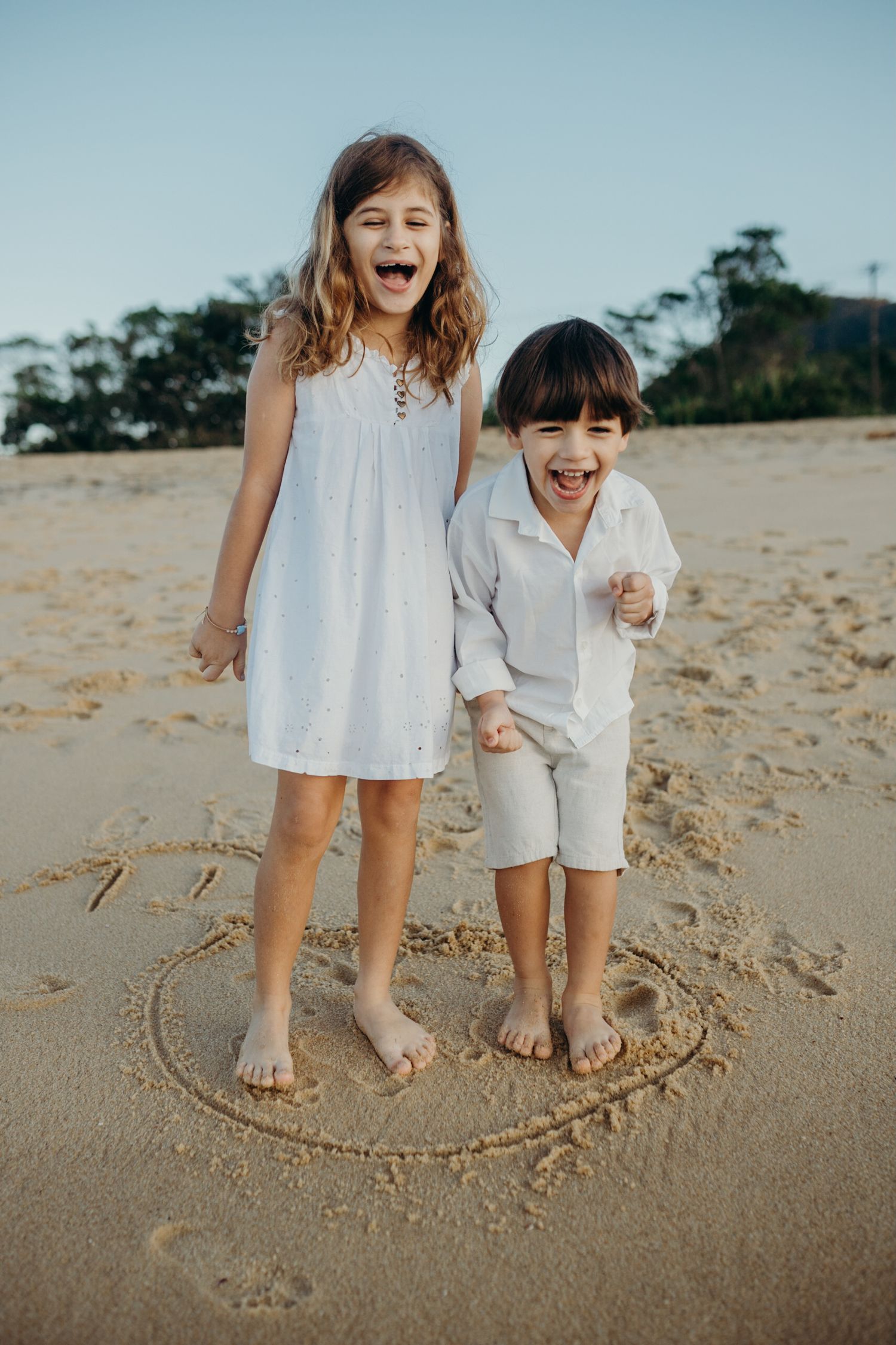 Children wearing white clothing laugh while standing in a heart drawn on sandy beach.