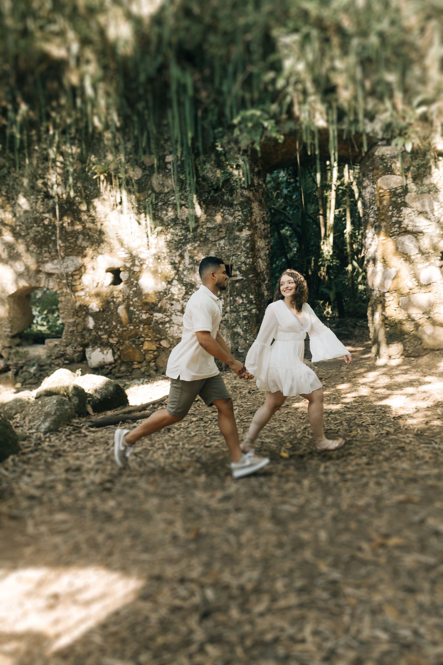 A playful couple runs through a scenic forest path in coordinated white outfits.