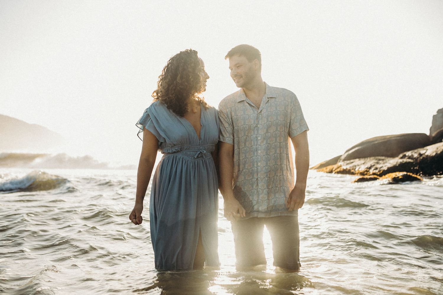 Couple wading in ocean water at sunset with rocky coastline in background.