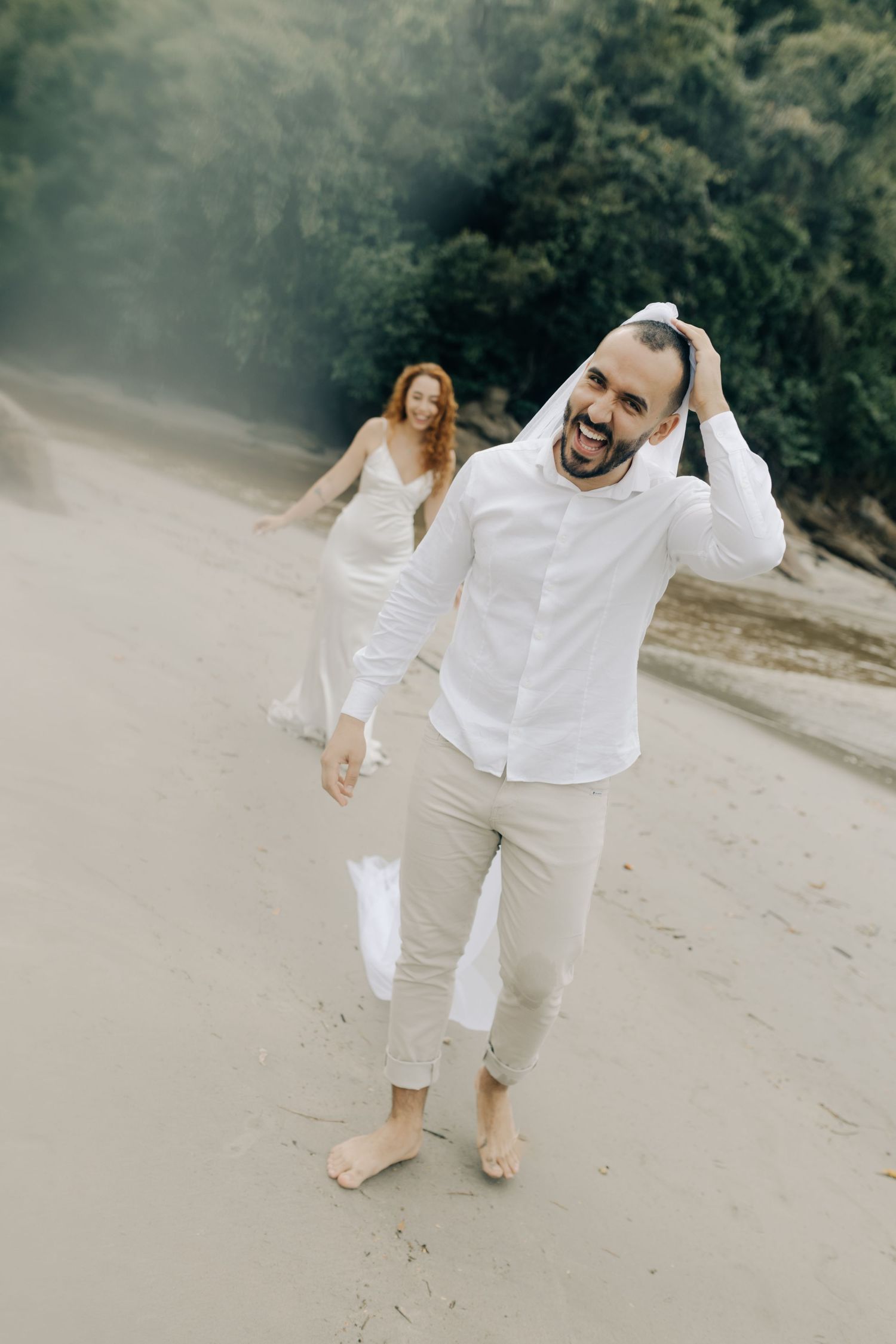 A couple dressed in white outfits dances playfully on a sandy beach.