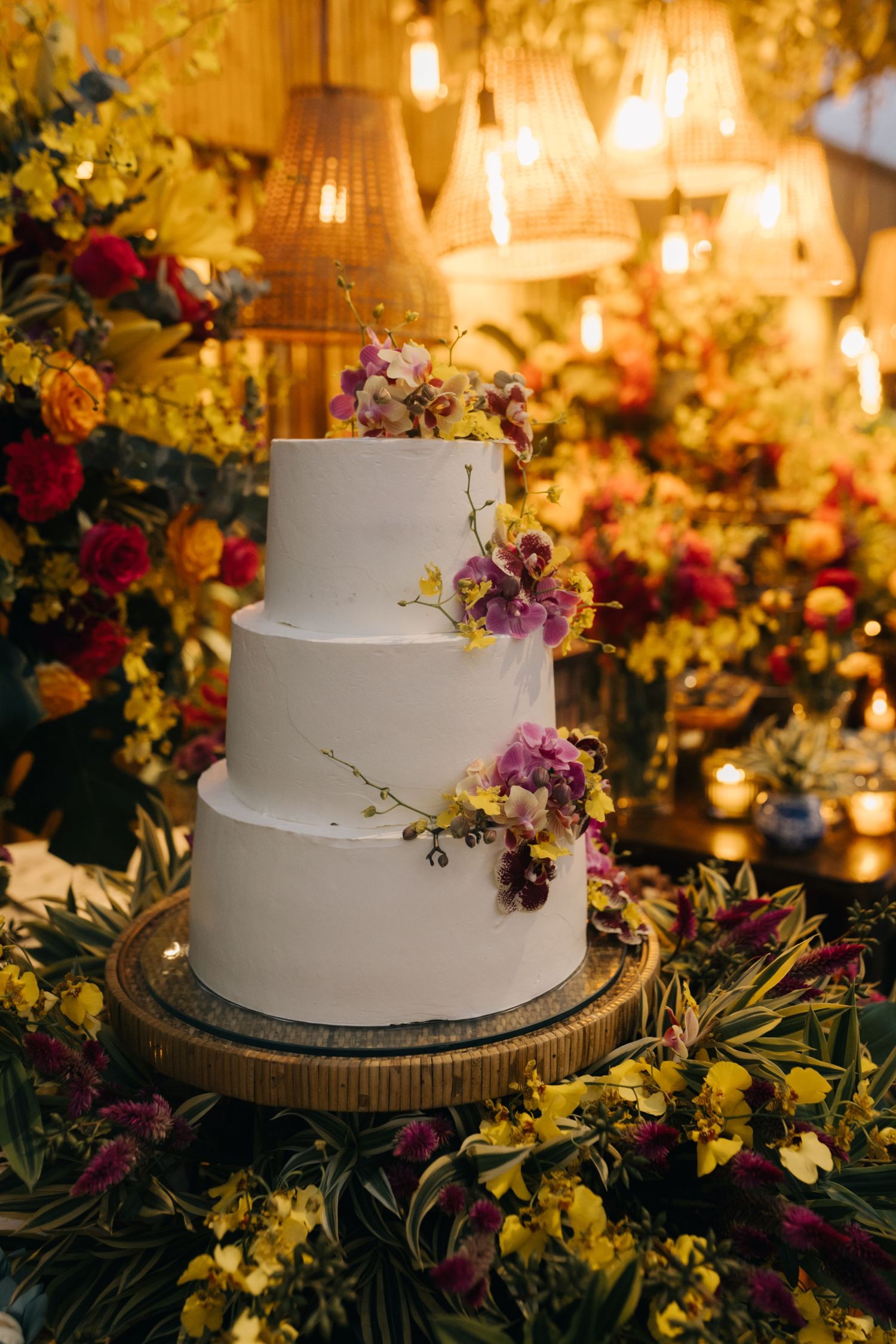 Three-tiered white wedding cake decorated with purple and pink flowers against a warmly lit floral backdrop.