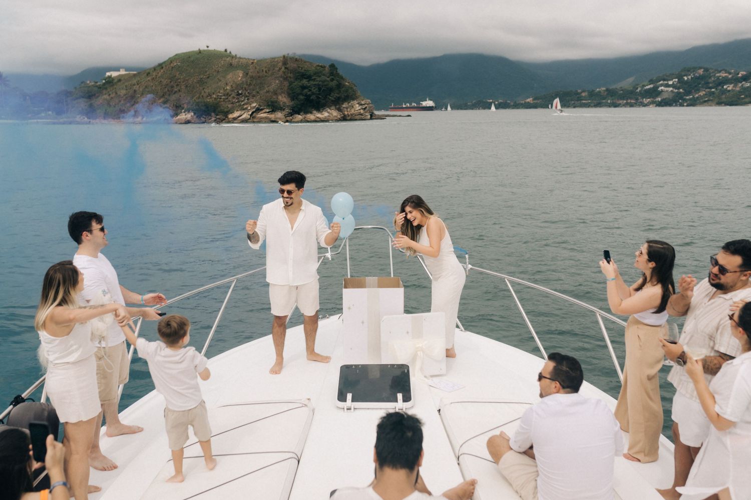 Guests in white and beige outfits celebrate on a yacht with mountains visible in the distance.