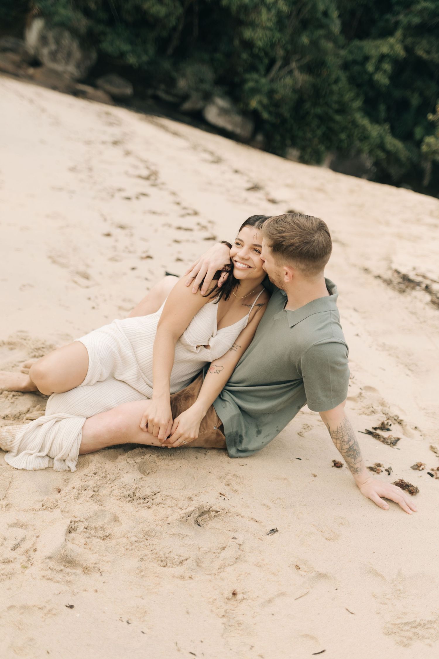 A couple relaxes on the sand during a romantic beach photoshoot near coastal rocks.