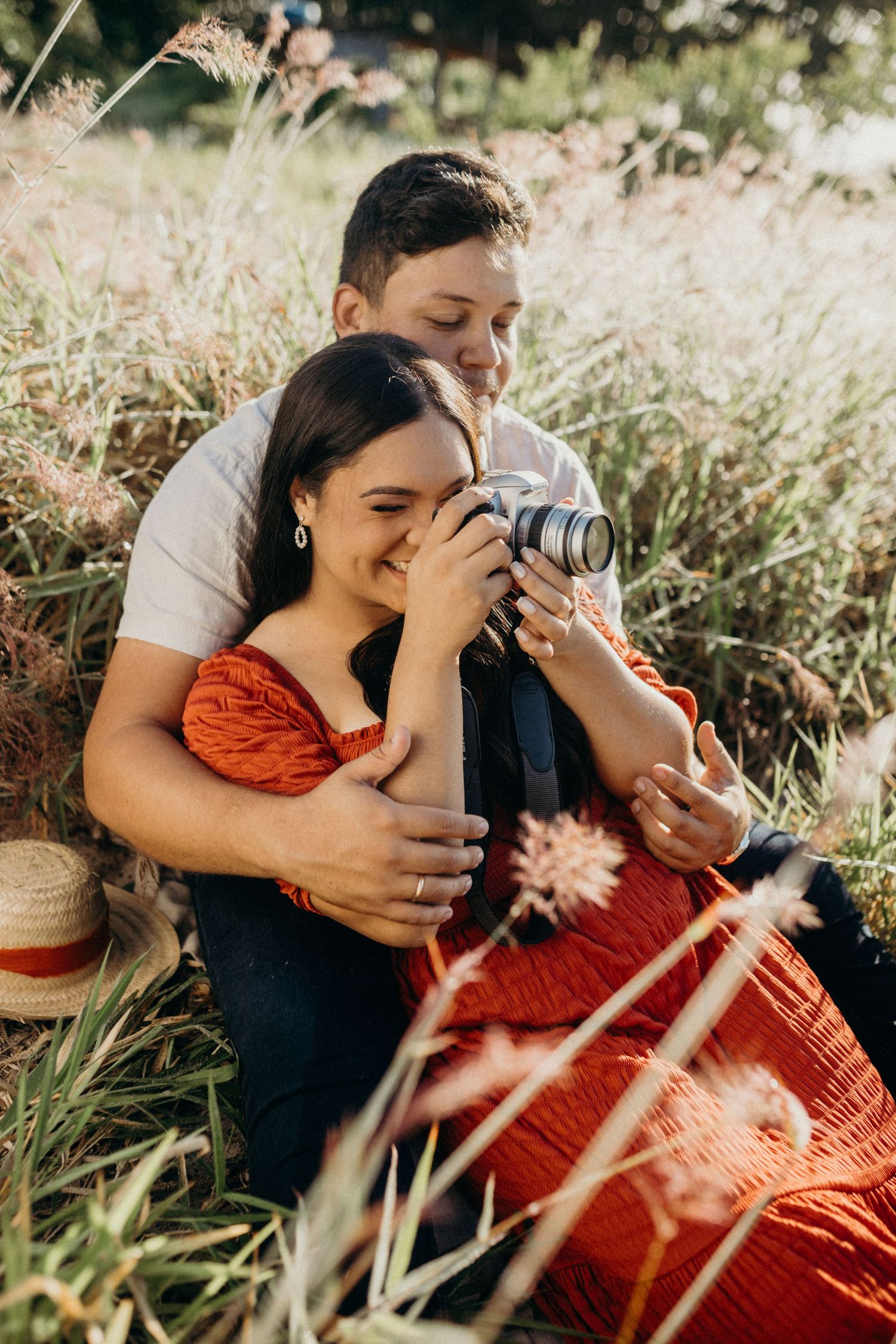Two people share tender moment while taking photos together among wild coastal grasses.