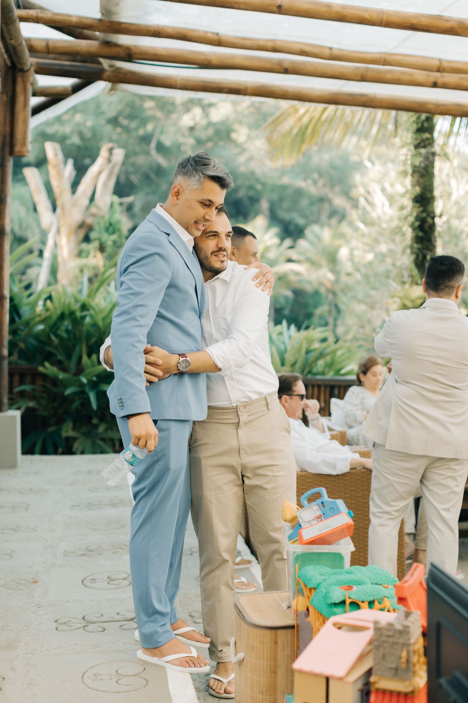 Two guests embrace at an outdoor wedding celebration wearing light colored suits and sneakers.
