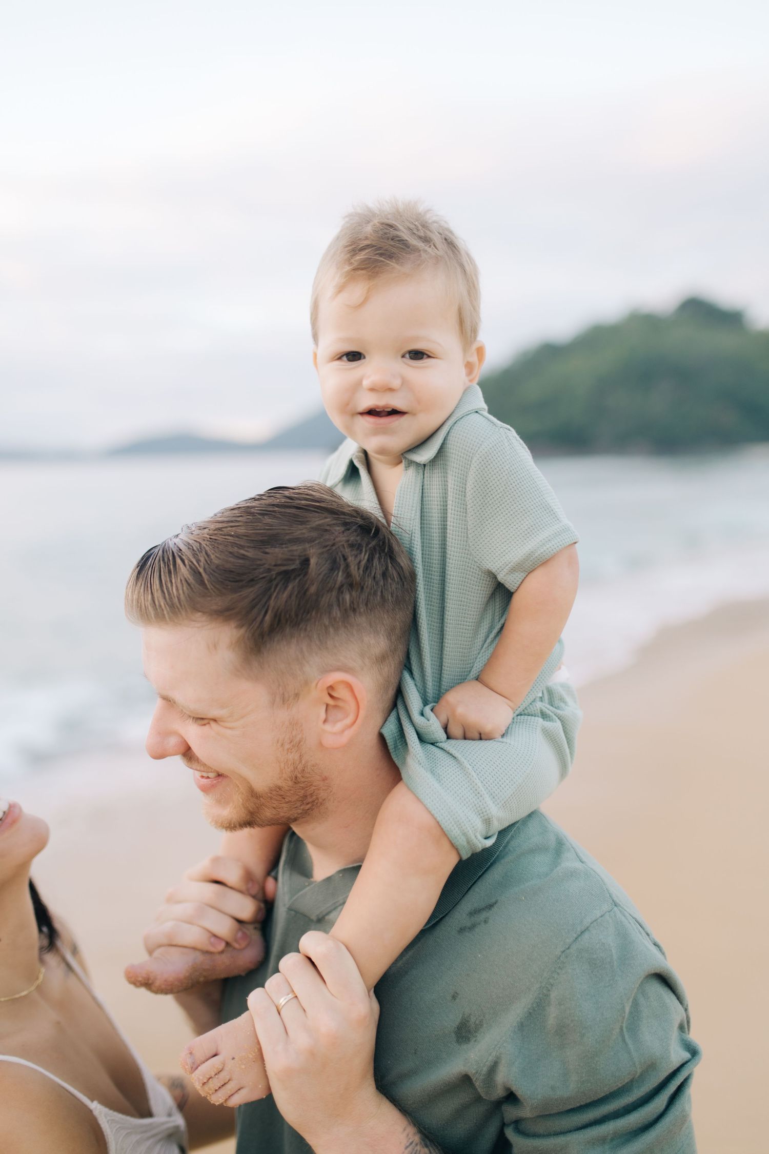 A toddler sits on someone's shoulders at the beach with ocean and hills in background.