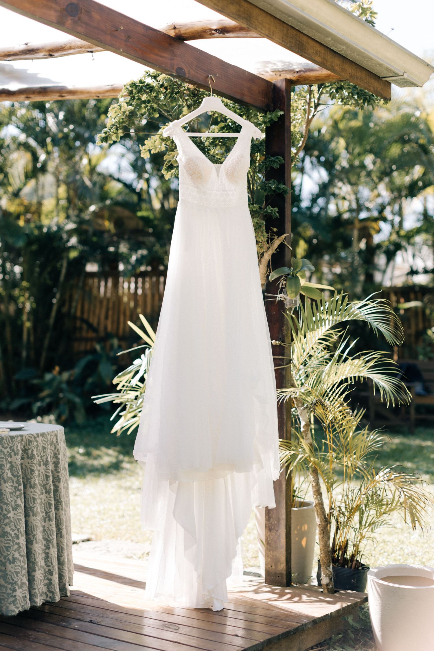 Elegant white wedding dress hanging from wooden beam in tropical garden setting.