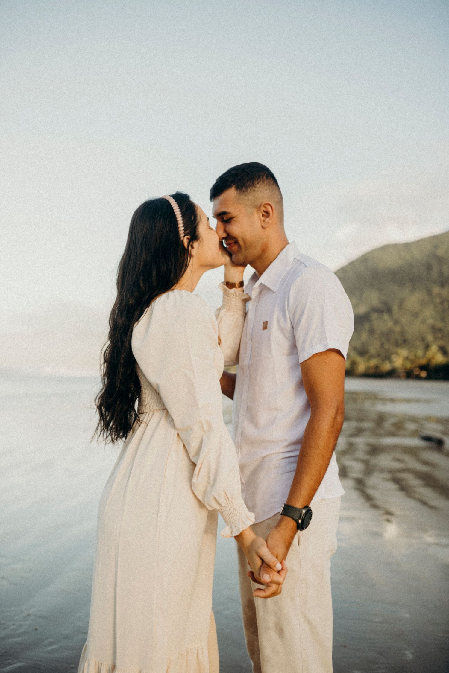 Romantic couple in light-colored outfits share an intimate moment at sunset on a beach with mountains in background.