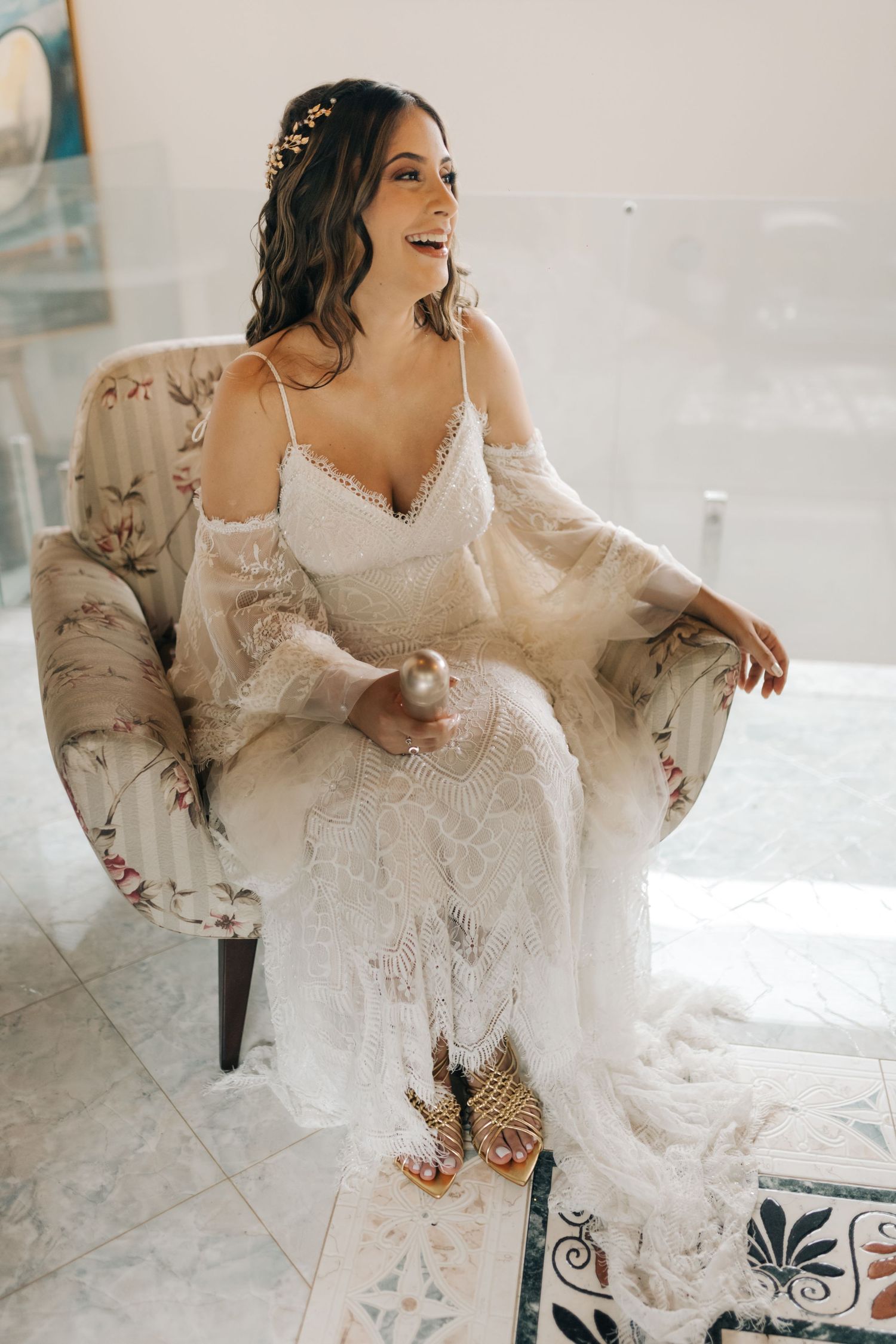 Candid moment of bride seated in floral chair wearing detailed lace gown.