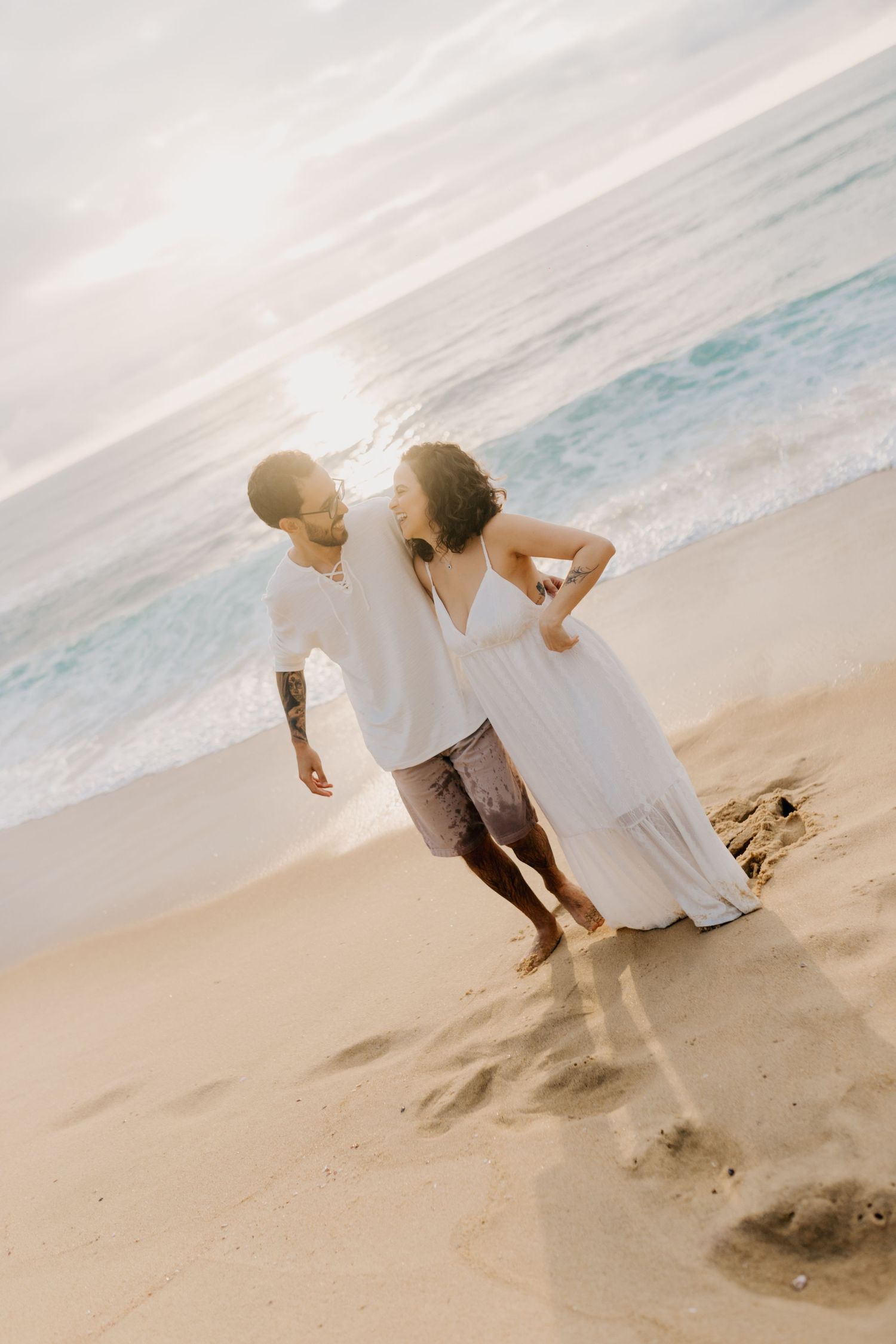 A couple in white outfits walks playfully along a beach with turquoise waves and sand in the background.
