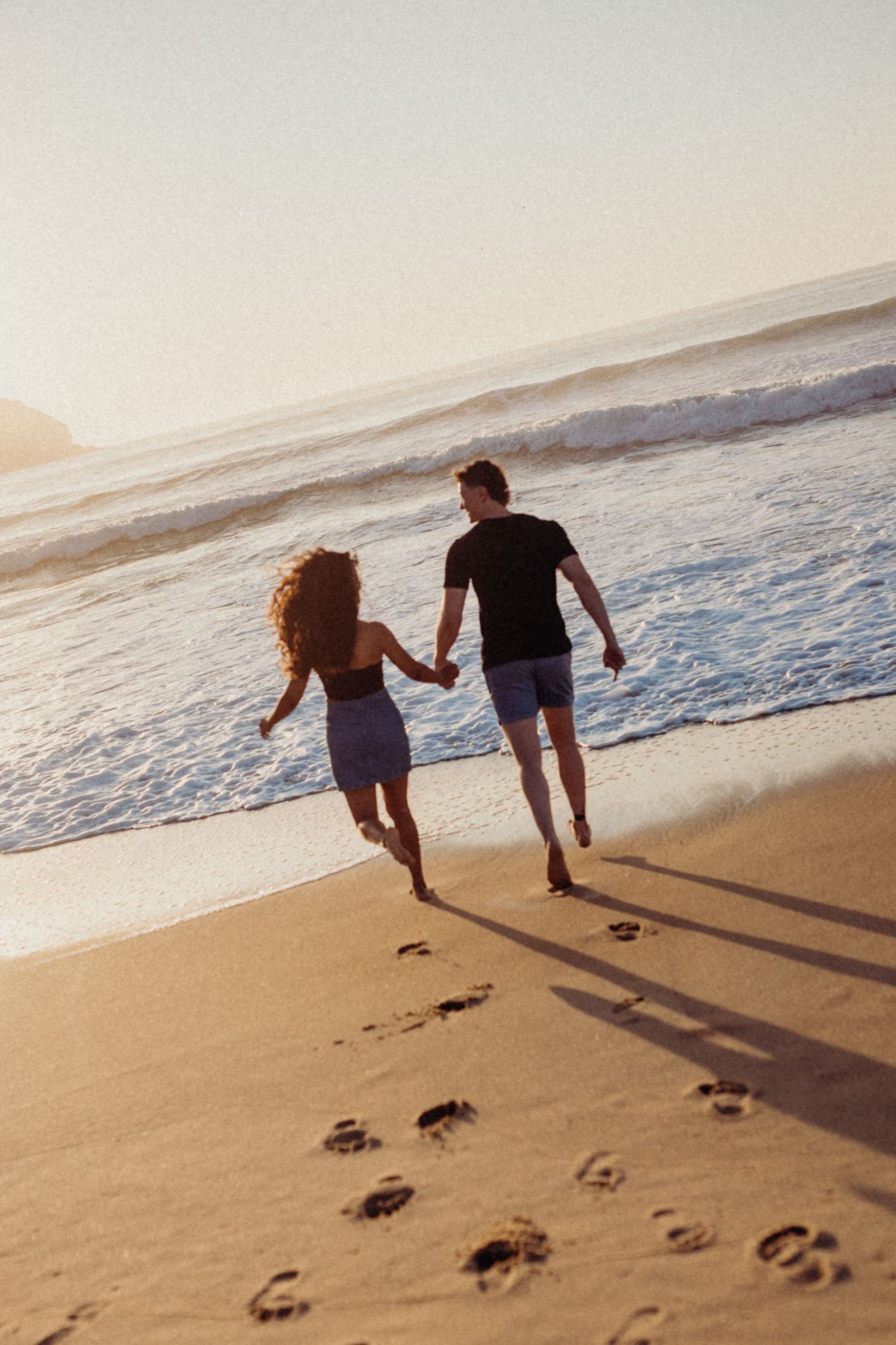 Couple runs playfully on beach leaving footprints in sand during sunset.