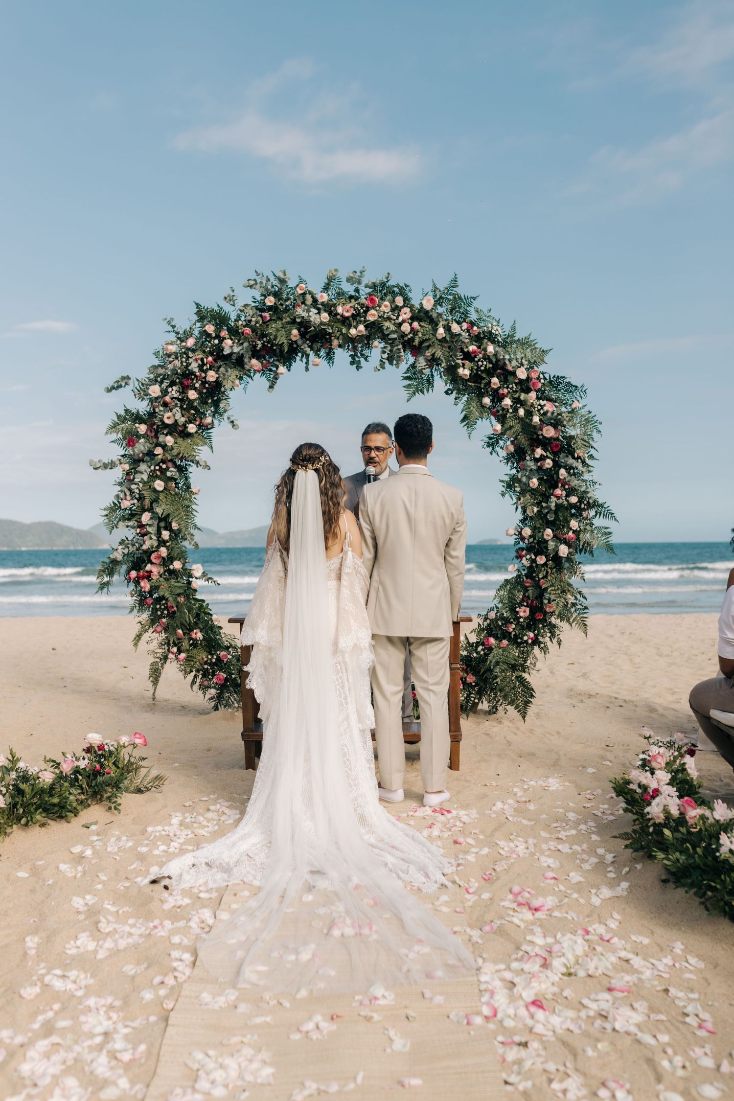 Floral wedding arch on beach with pink rose garlands and white fabric against ocean view.