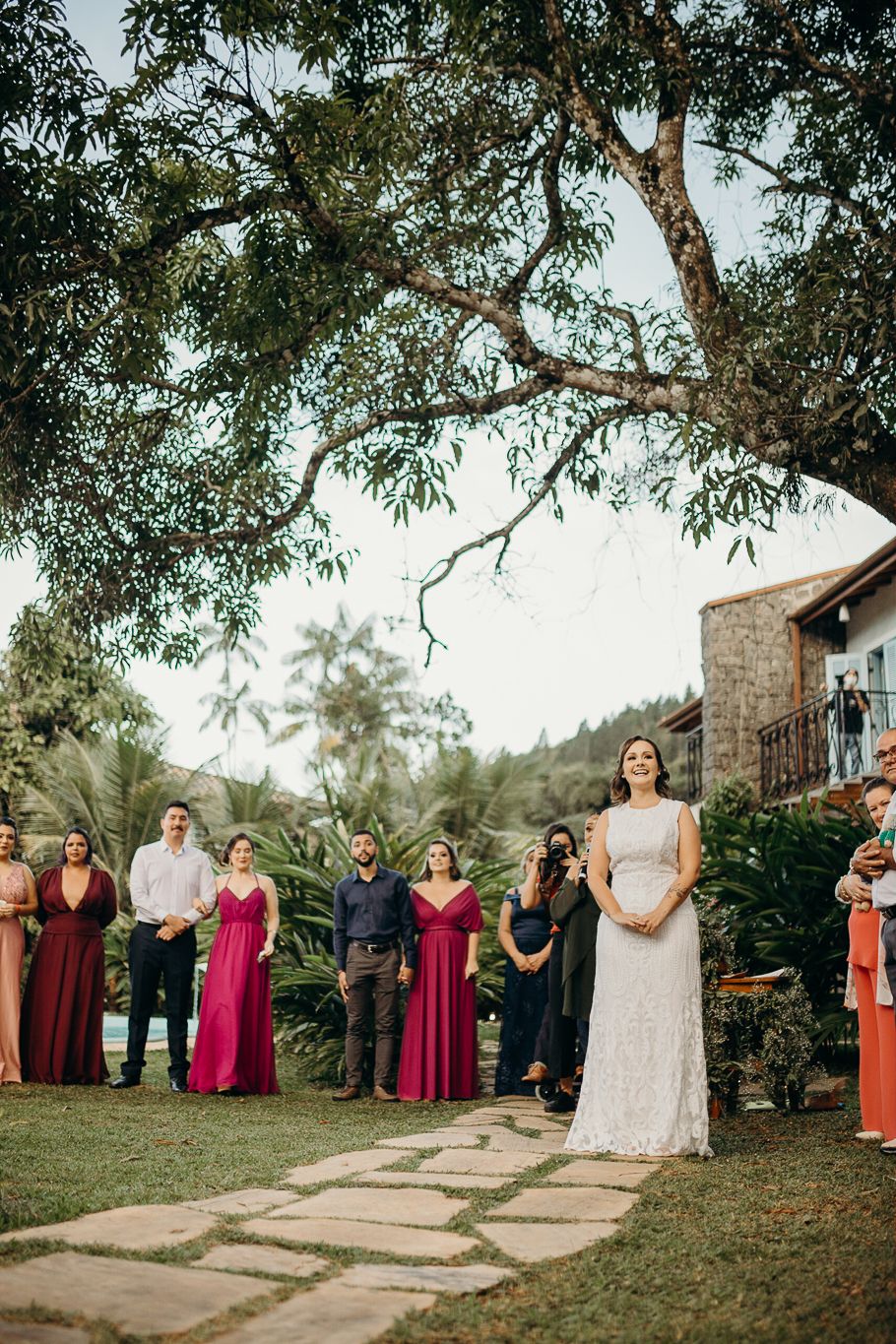 Wedding gathering on stone pathway with guests in formal attire and burgundy dresses under large tree branches.