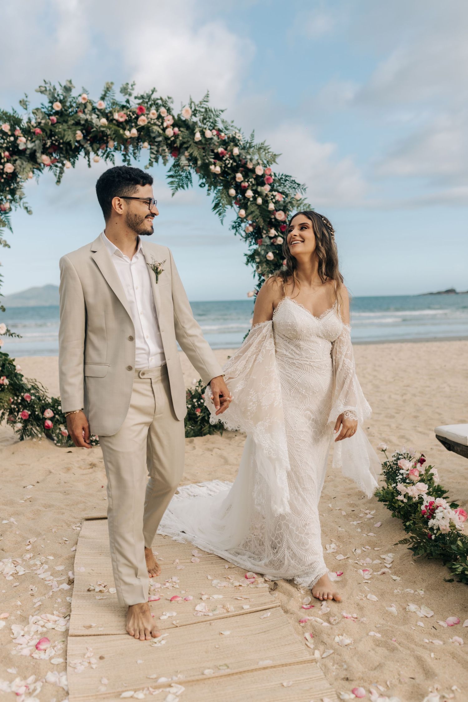 A beachside wedding ceremony with a floral arch and couple standing on a wooden walkway.