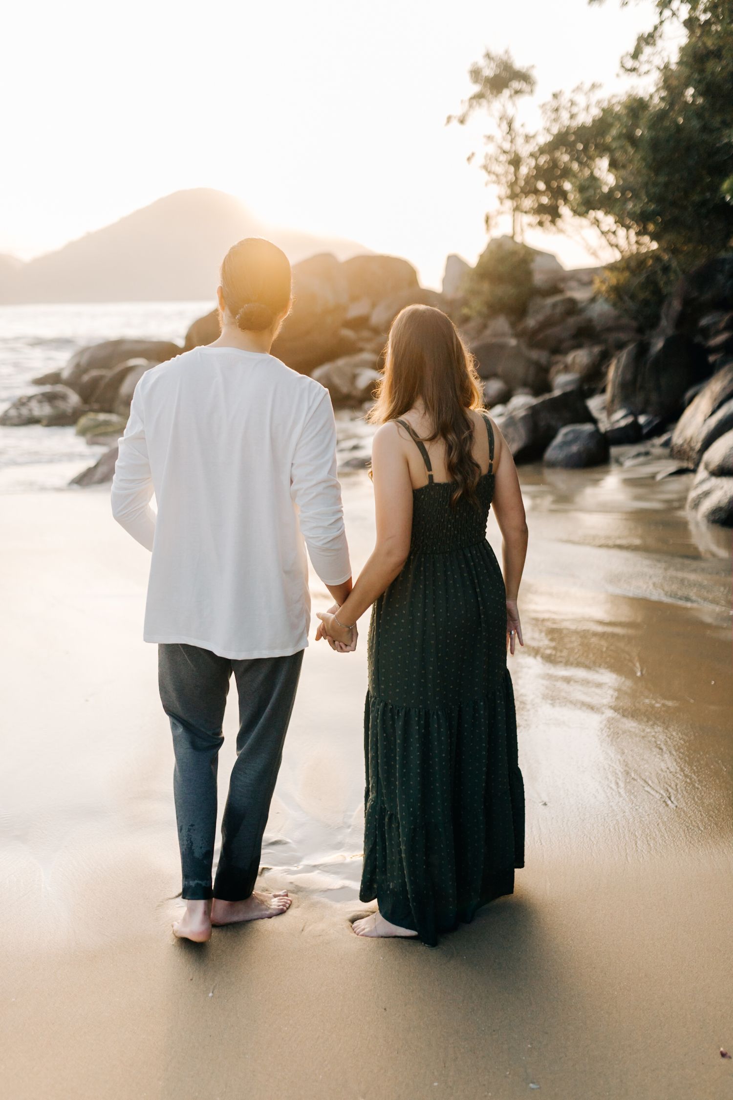 Couple holding hands while walking along shoreline during sunset with mountains and rocks in background.