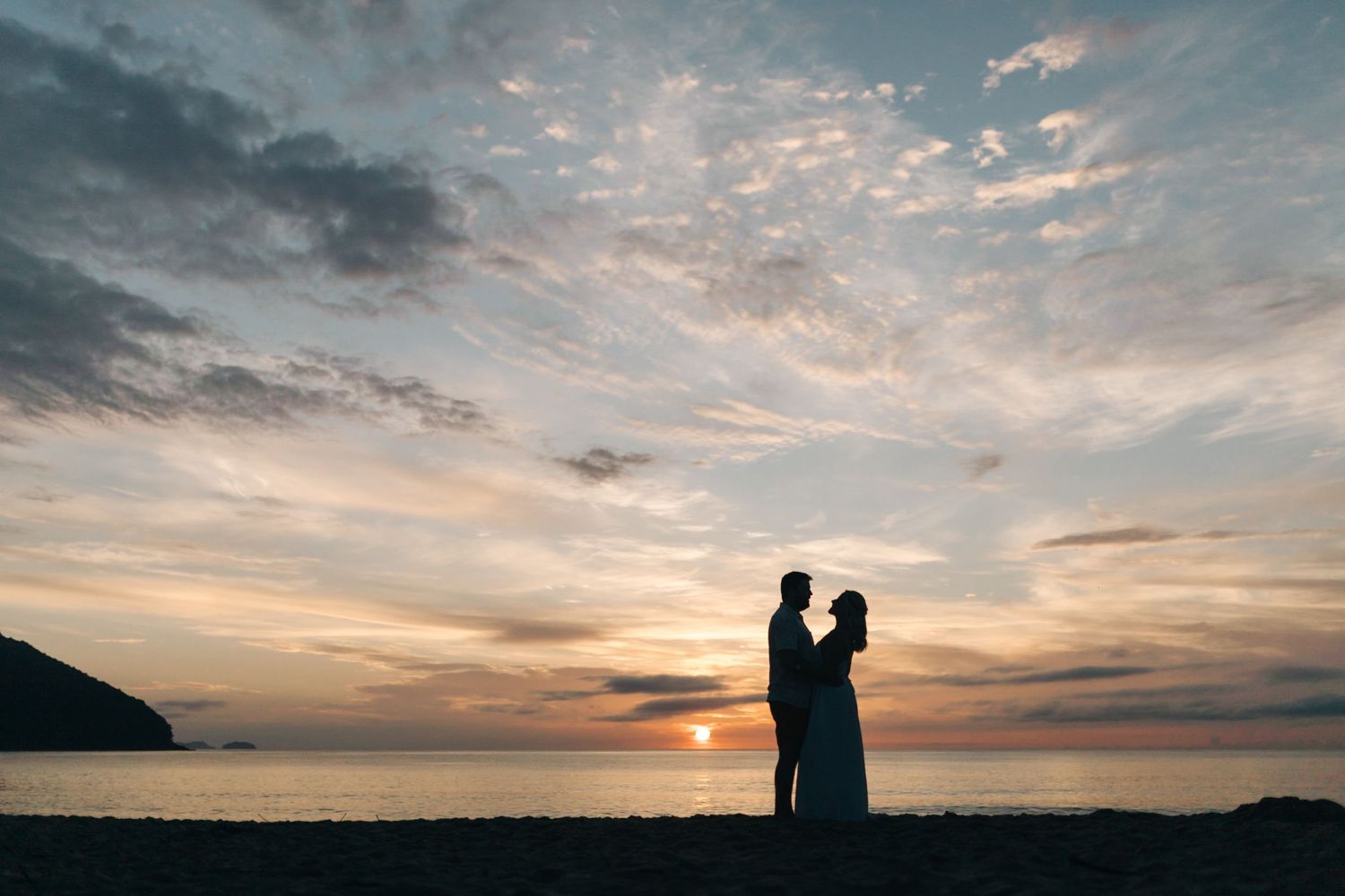 Two silhouettes stand together against a dramatic sunset sky at the beach.