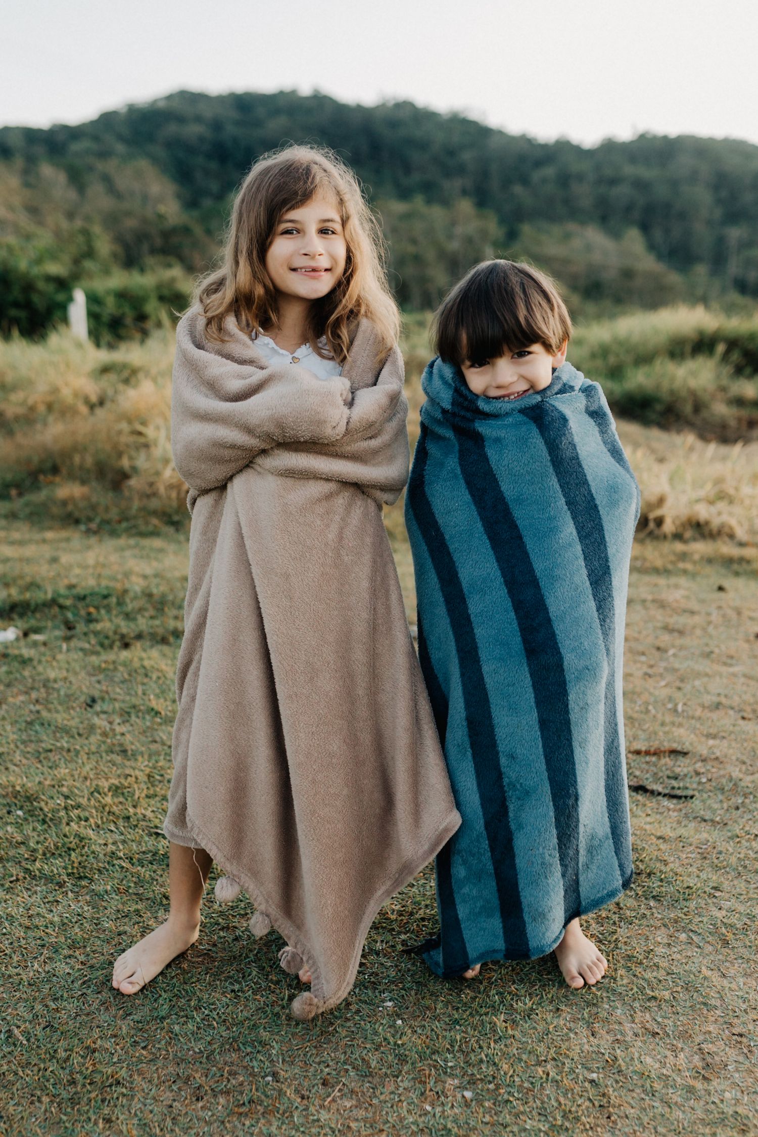 Two children wrapped in cozy blankets standing outdoors in natural setting.