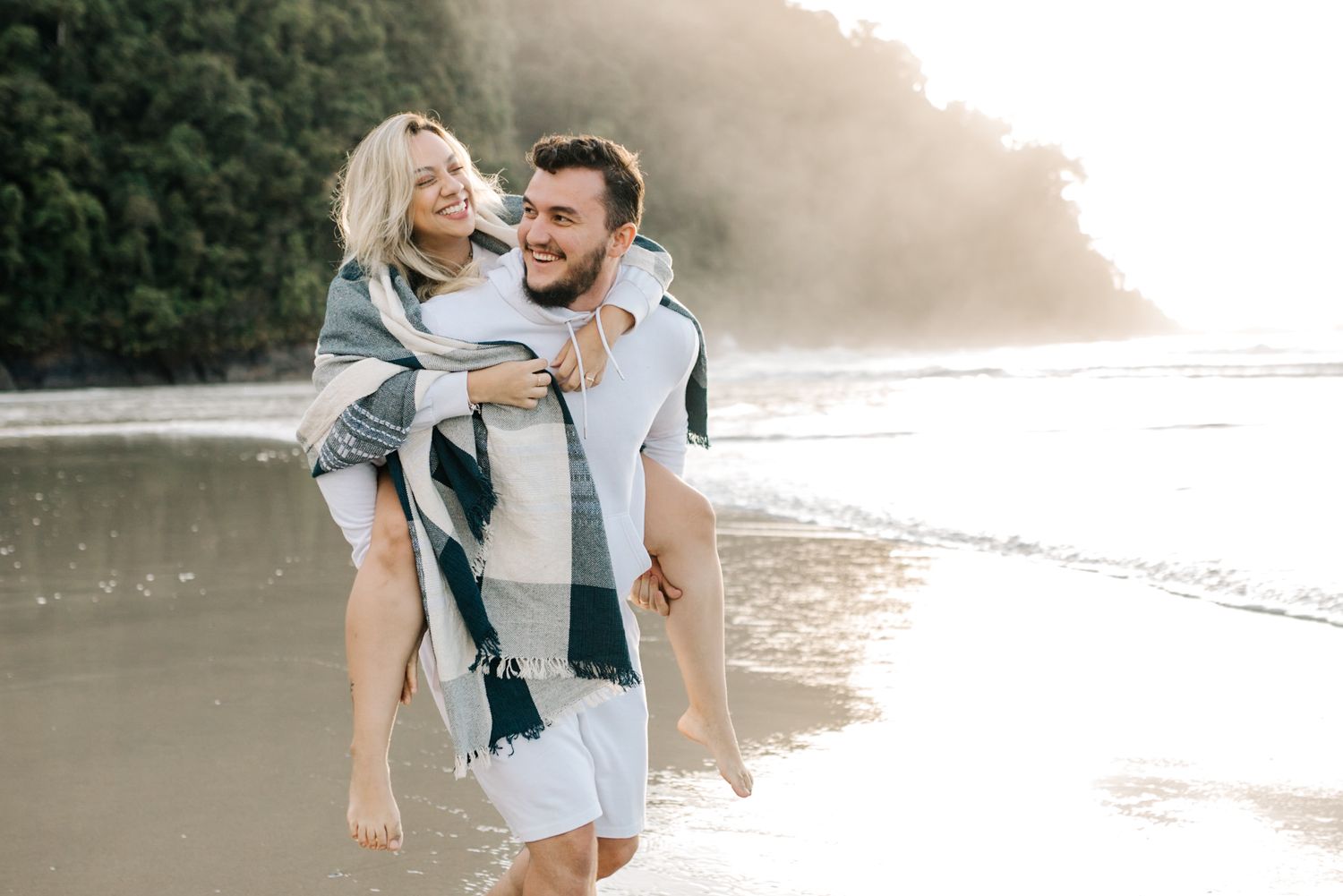 Joyful couple laughing during piggyback ride on sandy beach with forested cliffs in background.