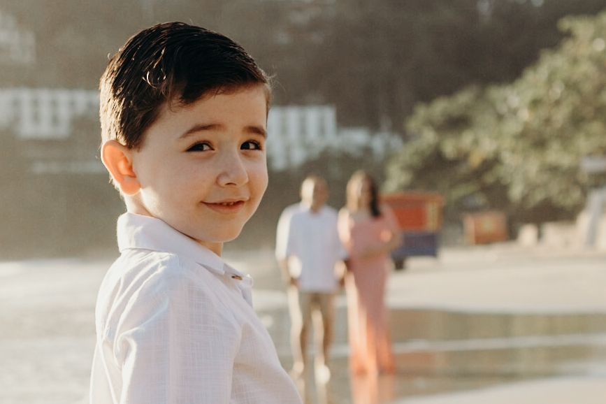 Young child smiling at camera on beach with blurred figures in background.